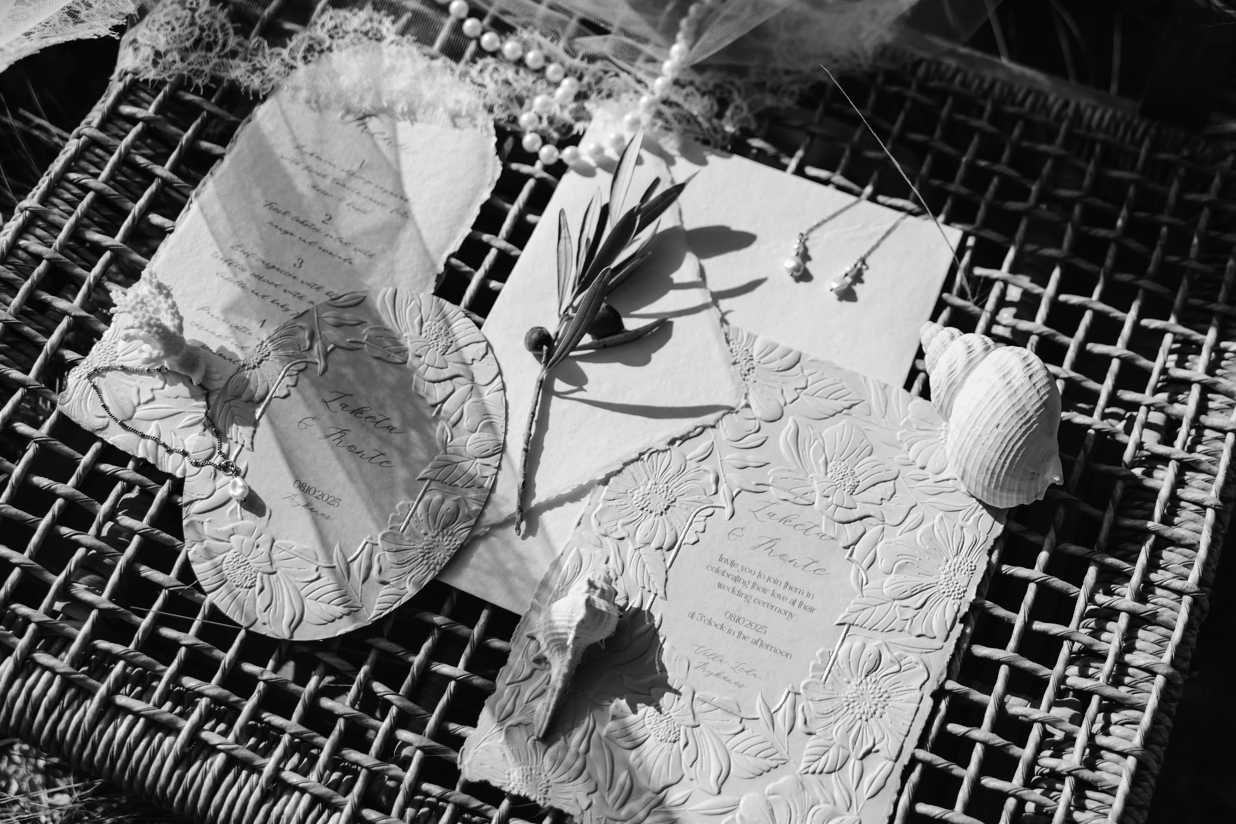 Wedding invitation with floral embossed design, seashells, and jewelry, placed on a woven surface.