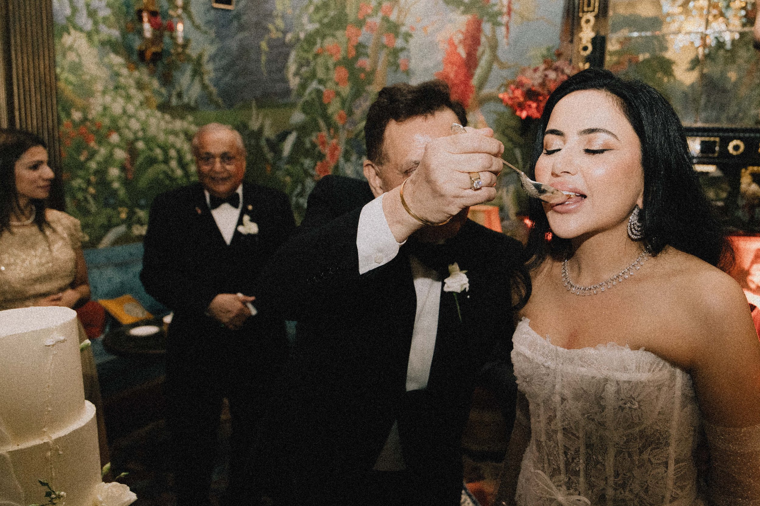A bride with dark hair and jewelry having her wedding cake fed to her by an older man at a wedding reception, with other guests in formal attire in the background.