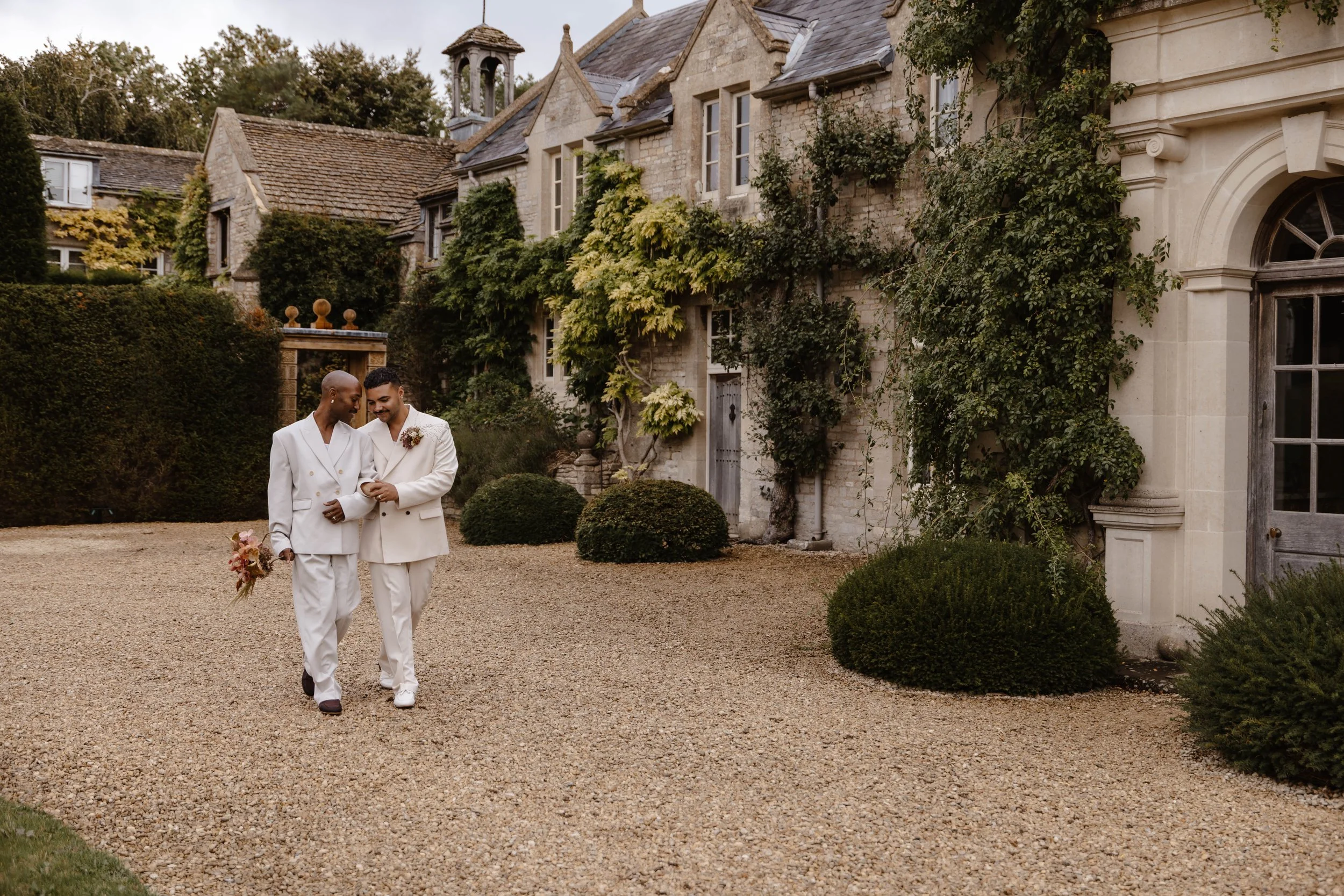 Two men in white suits walking arm in arm outside of a historic stone building with lush greenery in the background. beautiful LGBT wedding at Euridge manor 