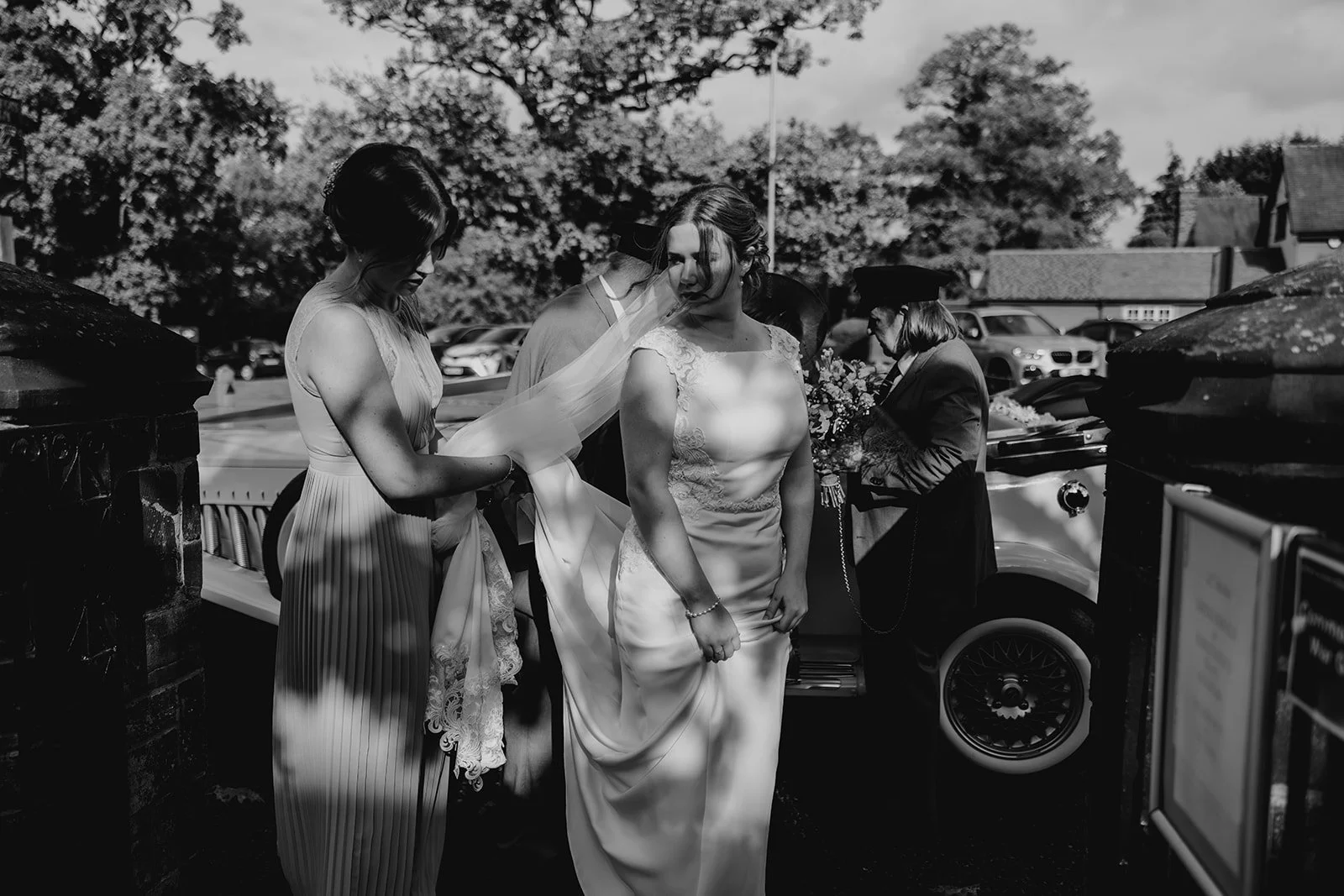 Black and white photo of a bride in a wedding dress standing outside near a vintage car, with women around her, some adjusting her dress and veil, and others holding flowers. wedding in cheshire, cheshire editorial wedding photographer 