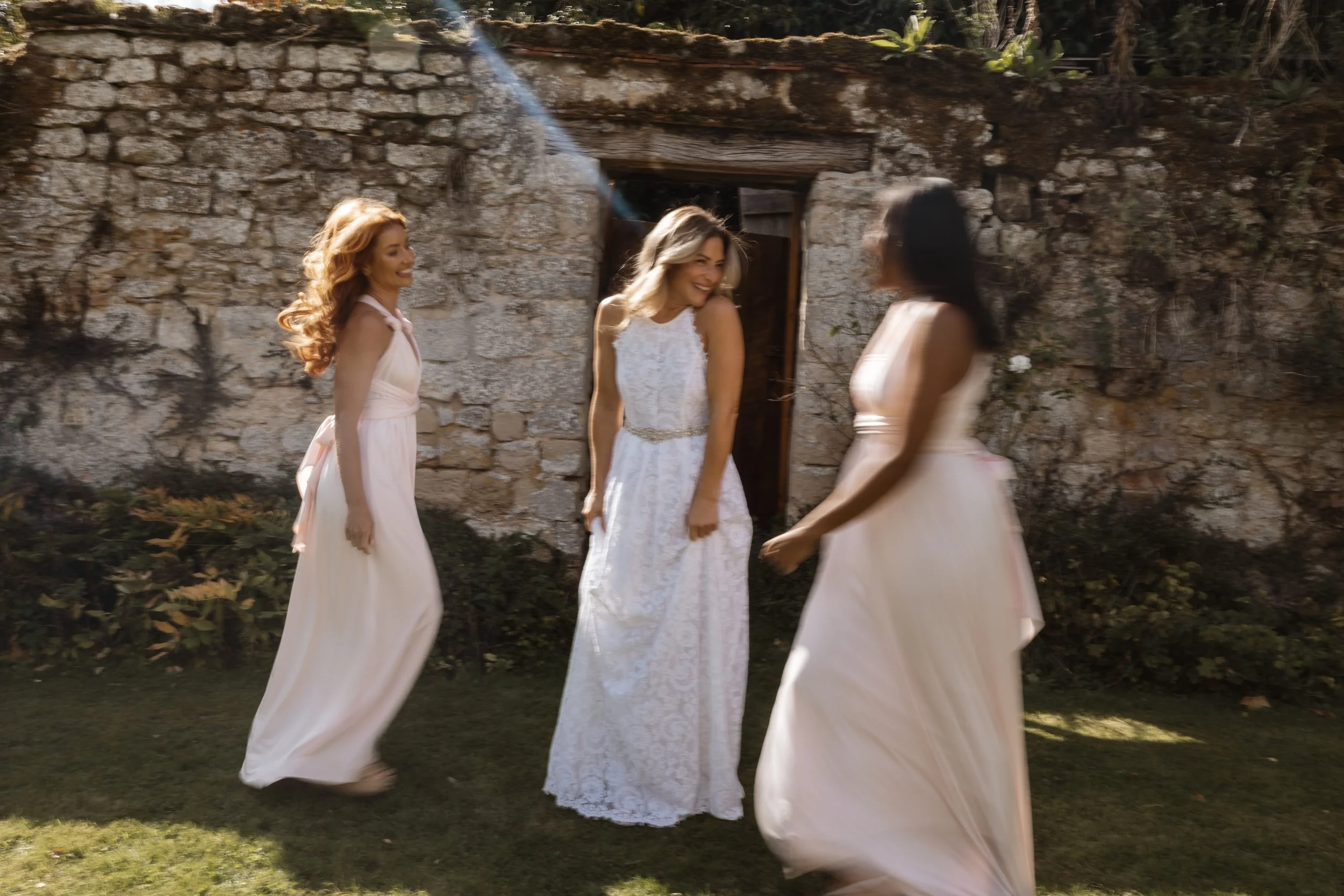 Four women in white dresses dancing and smiling outdoors in front of an old stone building. documentary and editorial wedding photographer