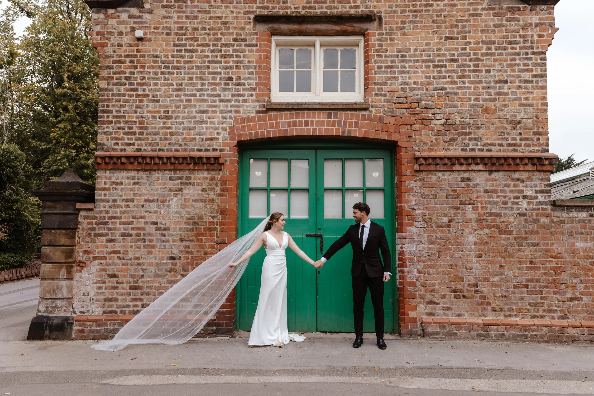 A bride and groom holding hands in front of a large green double door on a brick building. cheshire wedding venue, walton hall gardens weddings, documentary wedding photographer 