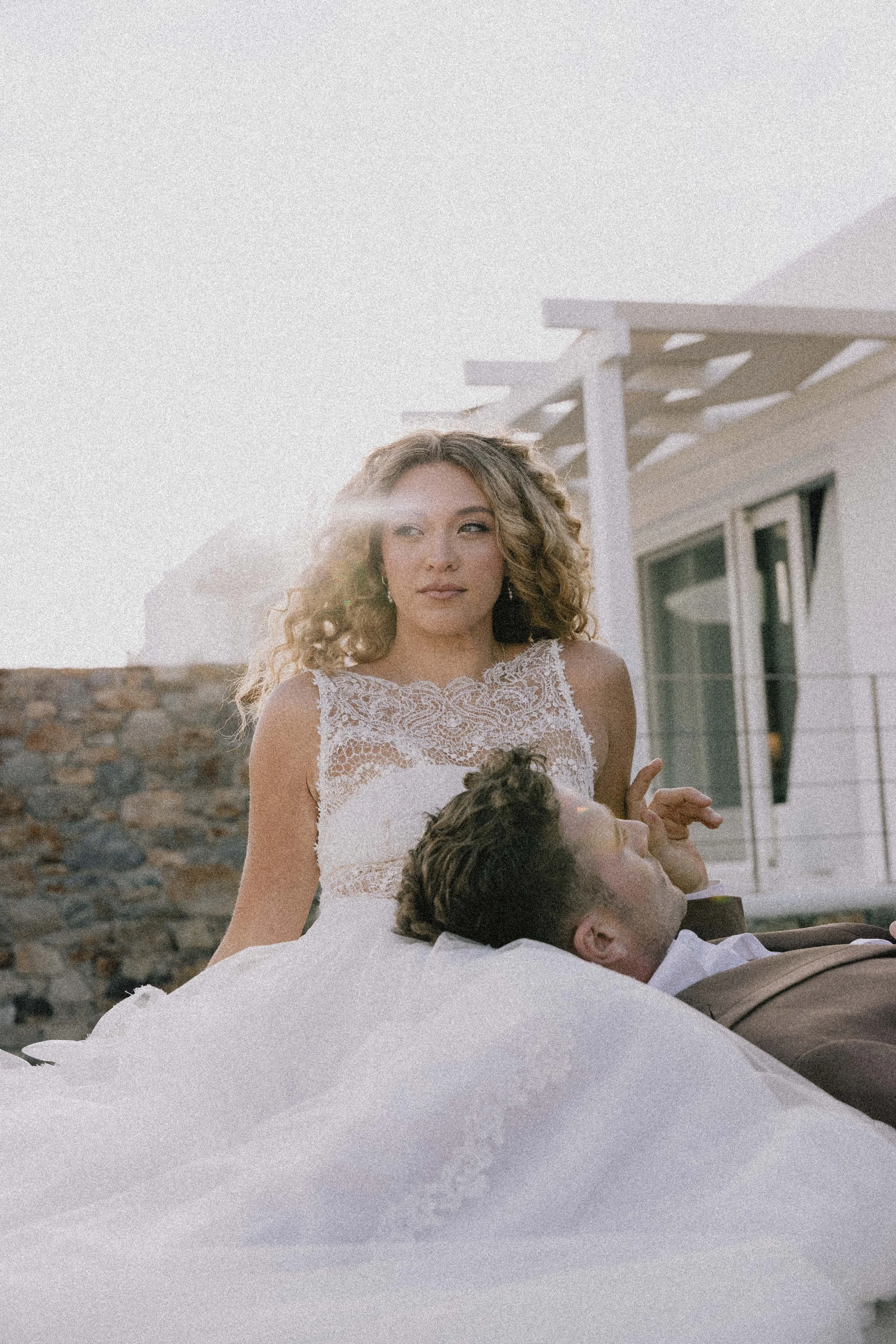 A woman in a white lace dress with curly hair stands behind a man in a white shirt and brown suit jacket who is lying down with his head on her lap, outside near a modern building with glass windows and a stone wall.