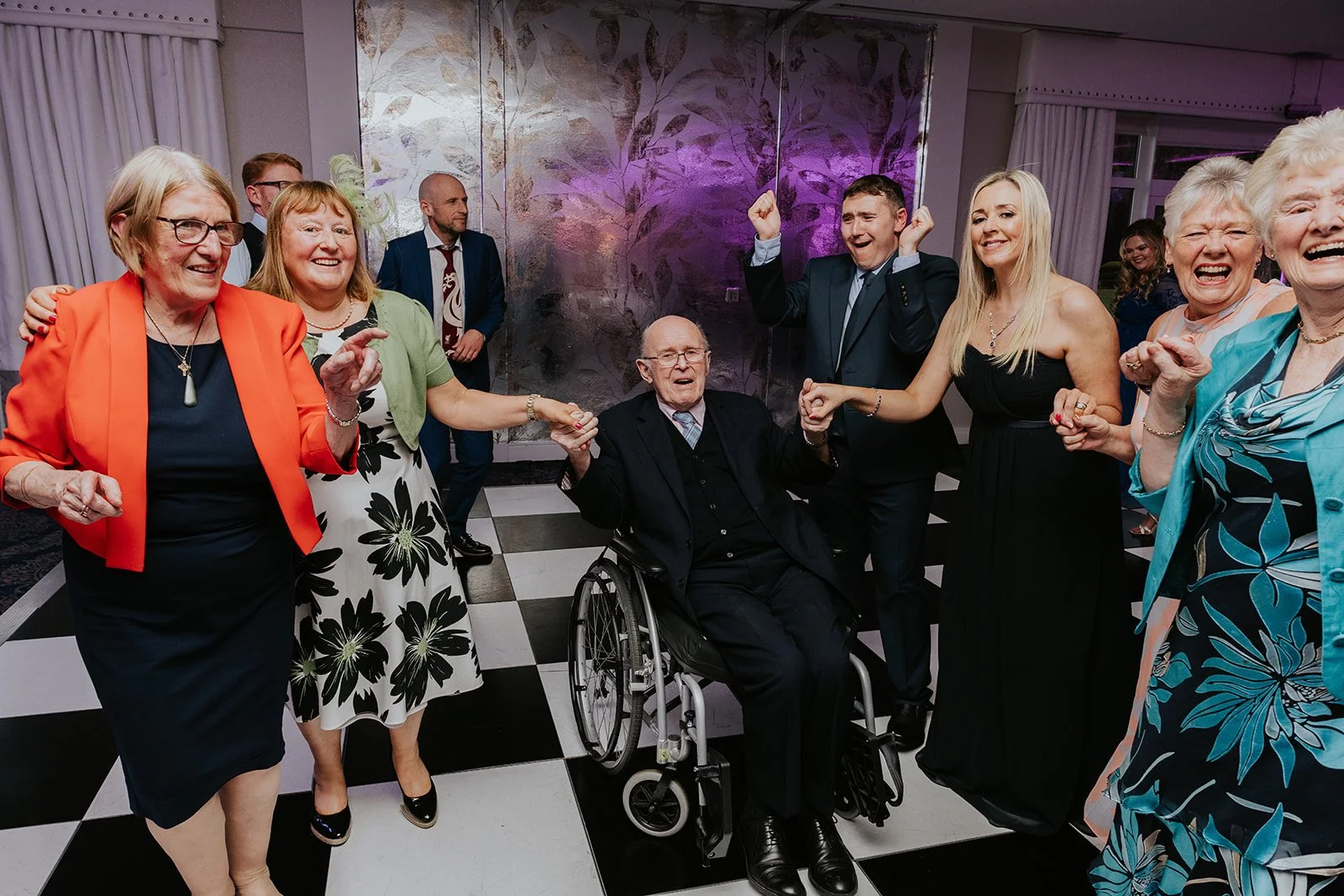 A group of elderly and young people dancing and celebrating together at a social event, with a man in a wheelchair in the center holding hands with two women, and others smiling and dancing around him. Merrydale Manor Wedding Venue