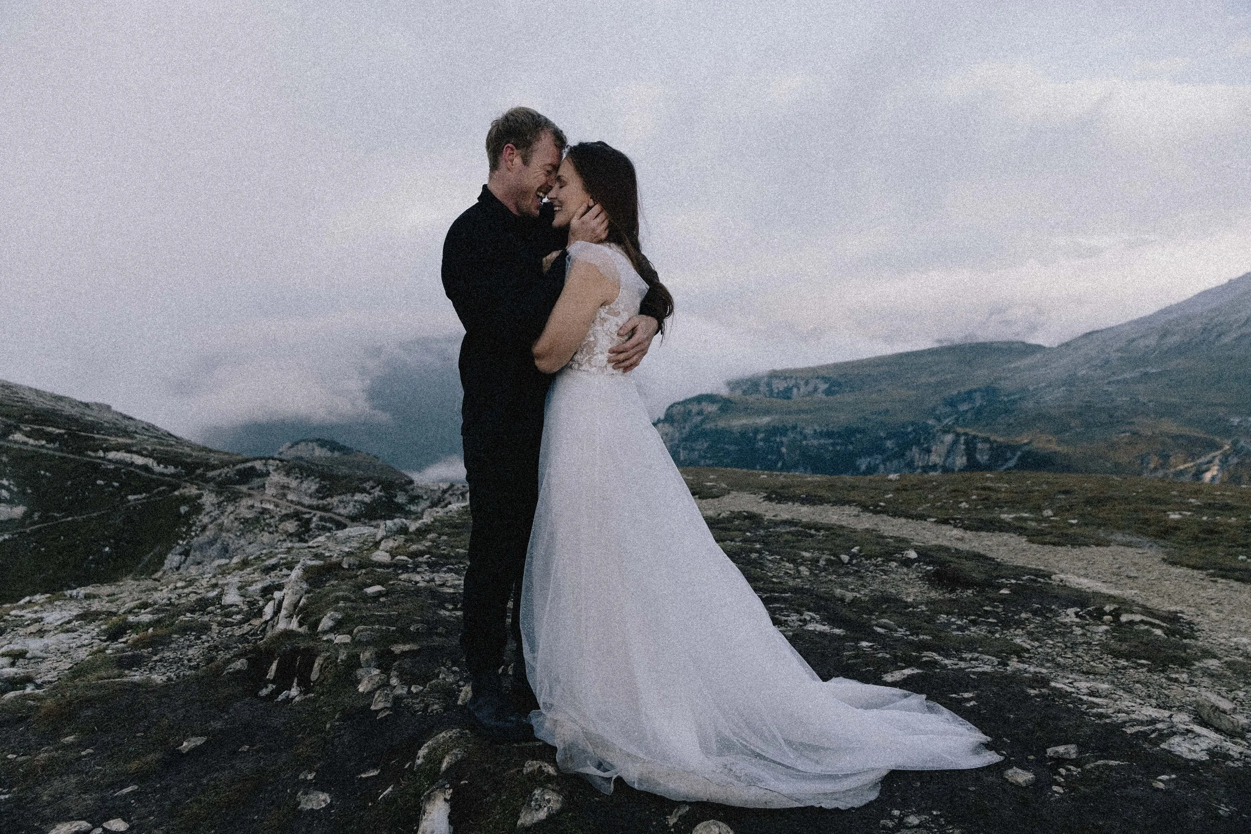 A bride and groom embrace on a rocky mountainous landscape during overcast weather, the bride in a white wedding gown and the groom in a black suit.