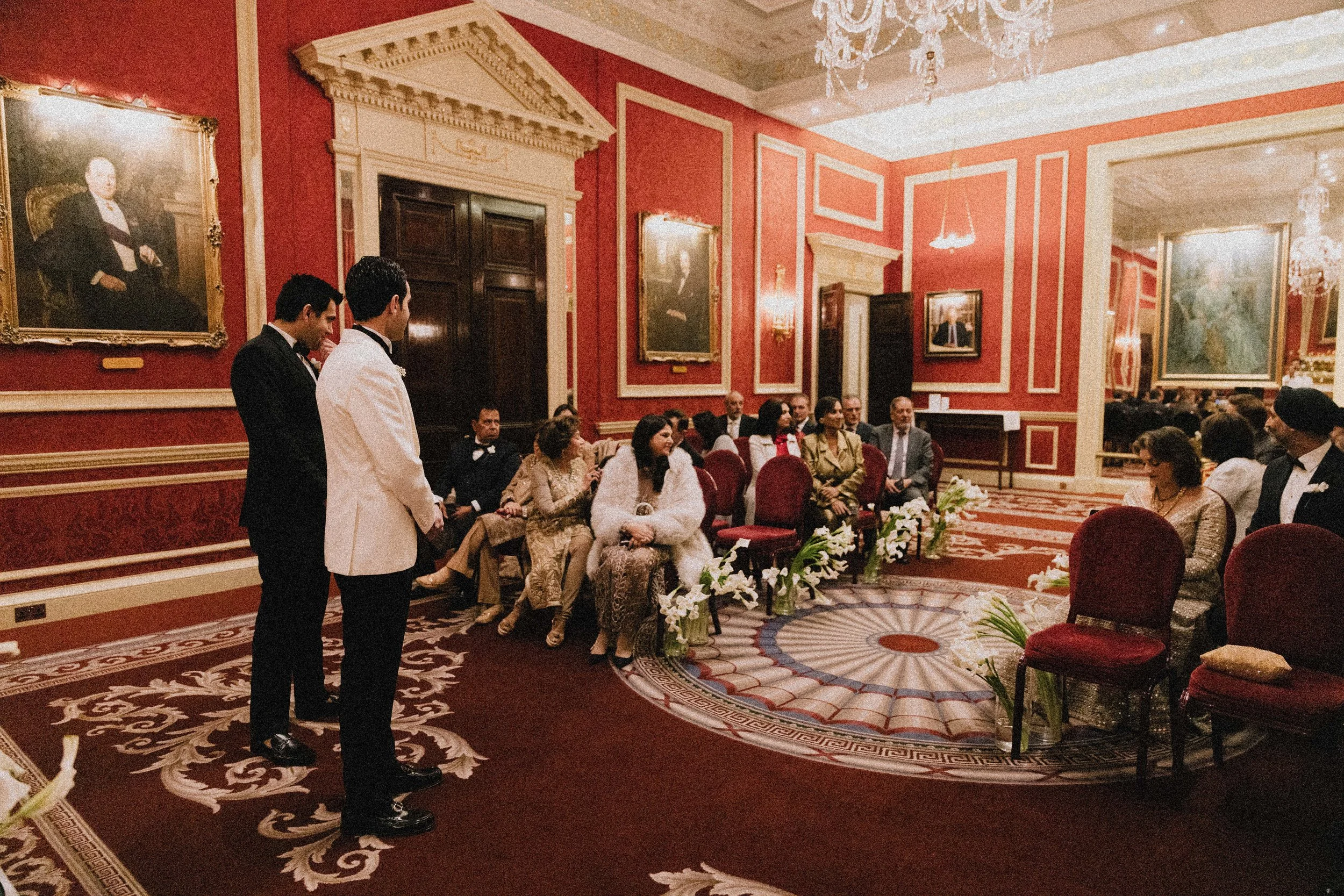  Several guests are seated on red chairs, and two men are standing in front of the seated guests, possibly during a speech or presentation. London wedding photography RAC wedding venue 
