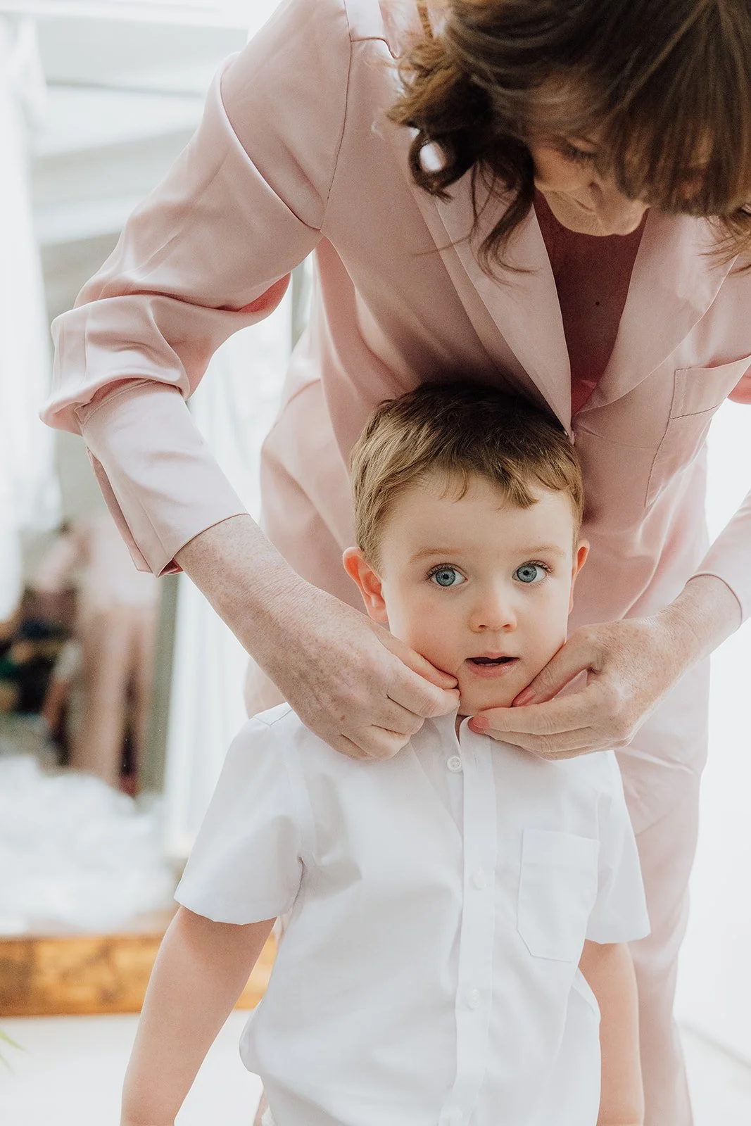 A woman in a pink silk robe is helping a young boy in a white shirt with a costume or outfit, adjusting his collar and neck. The boy has blue eyes and light brown hair, and appears to be looking at the camera. wedding at styal lodge