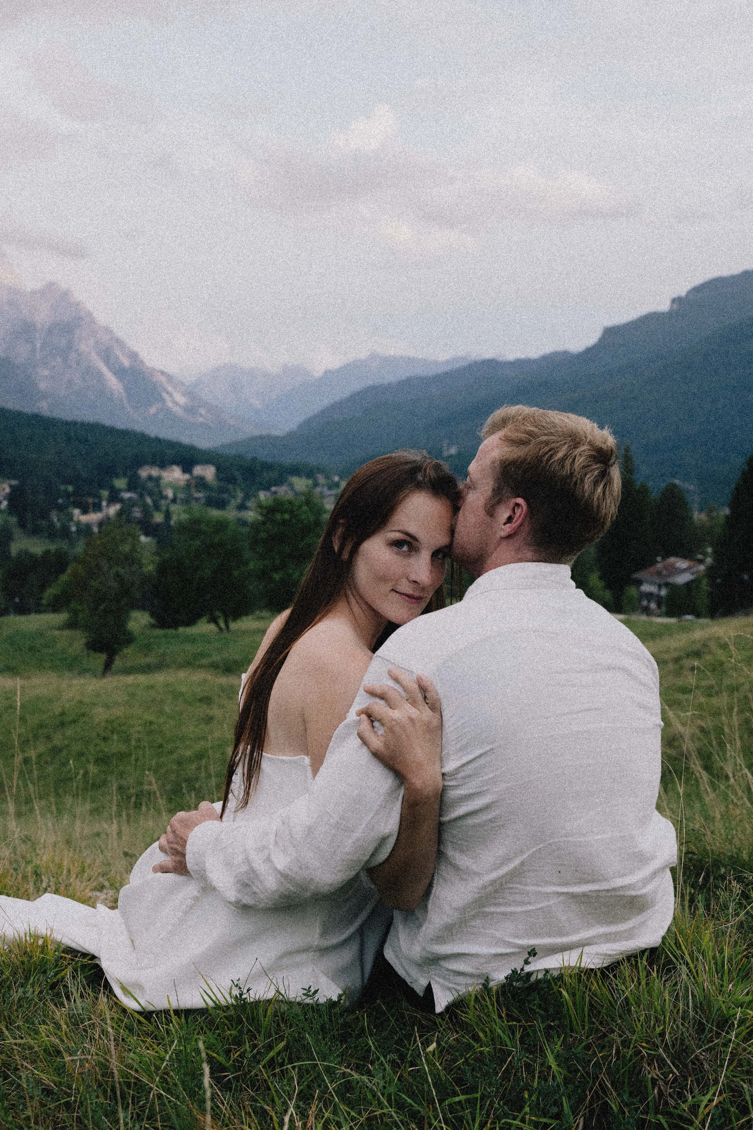 A couple sits close together on the grass in a scenic mountain landscape during dusk, with the man kissing the woman's forehead. wedding in italy 