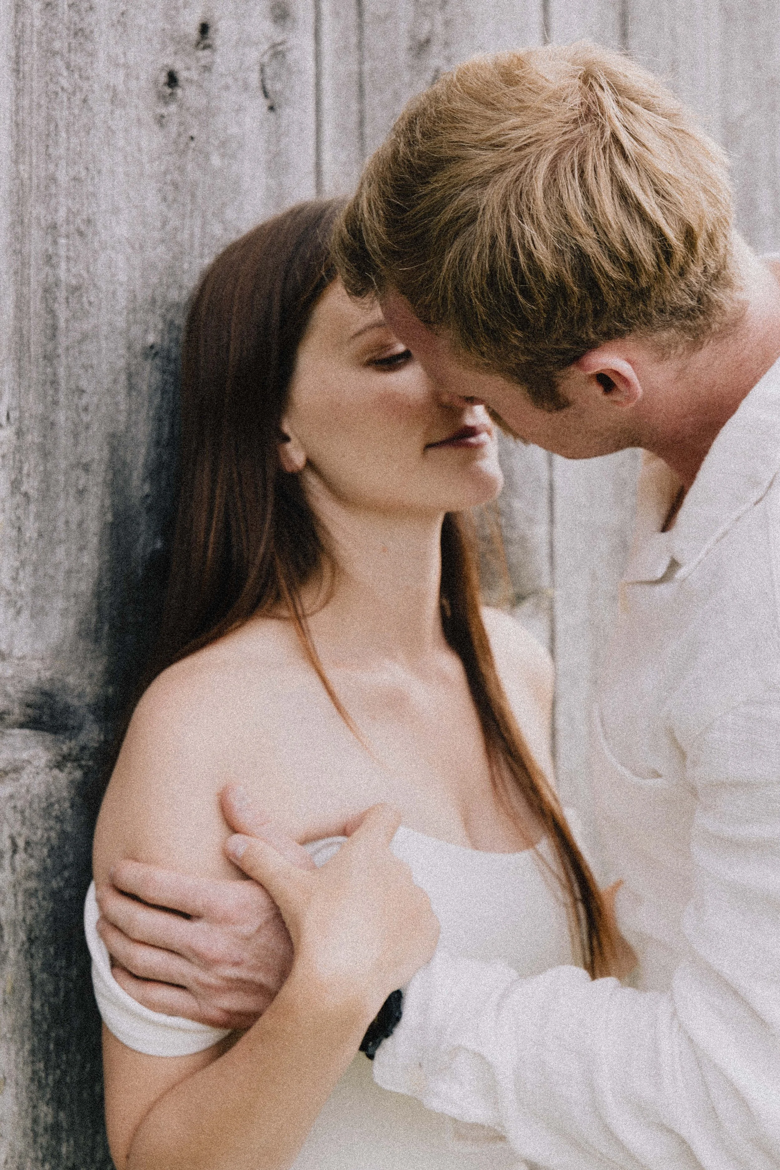A man and woman are close to each other, about to kiss, against a wooden background. The woman has long brown hair and is wearing a white off-shoulder top. The man has short blonde hair and is wearing a white shirt.