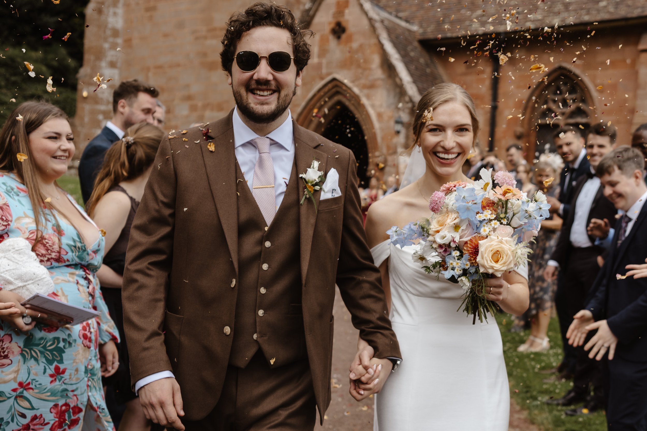 A happy bride and groom walk hand in hand after their wedding ceremony outside a church, surrounded by friends and family celebrating with confetti.