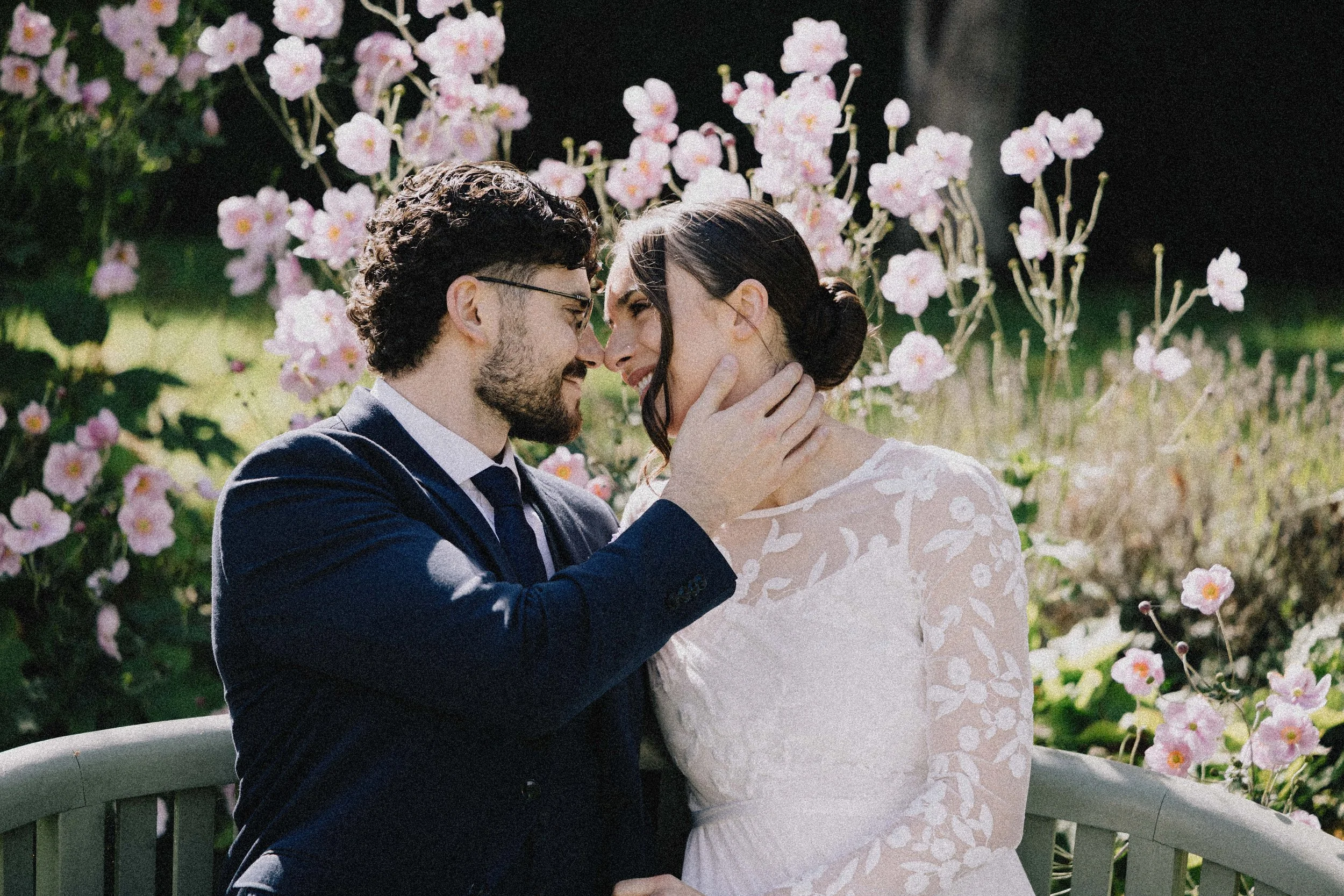 A couple dressed in wedding attire sitting on a bench, gently touching noses and smiling in a garden with pink flowers in the background. a 35mm film photo of wedding at rowton hall