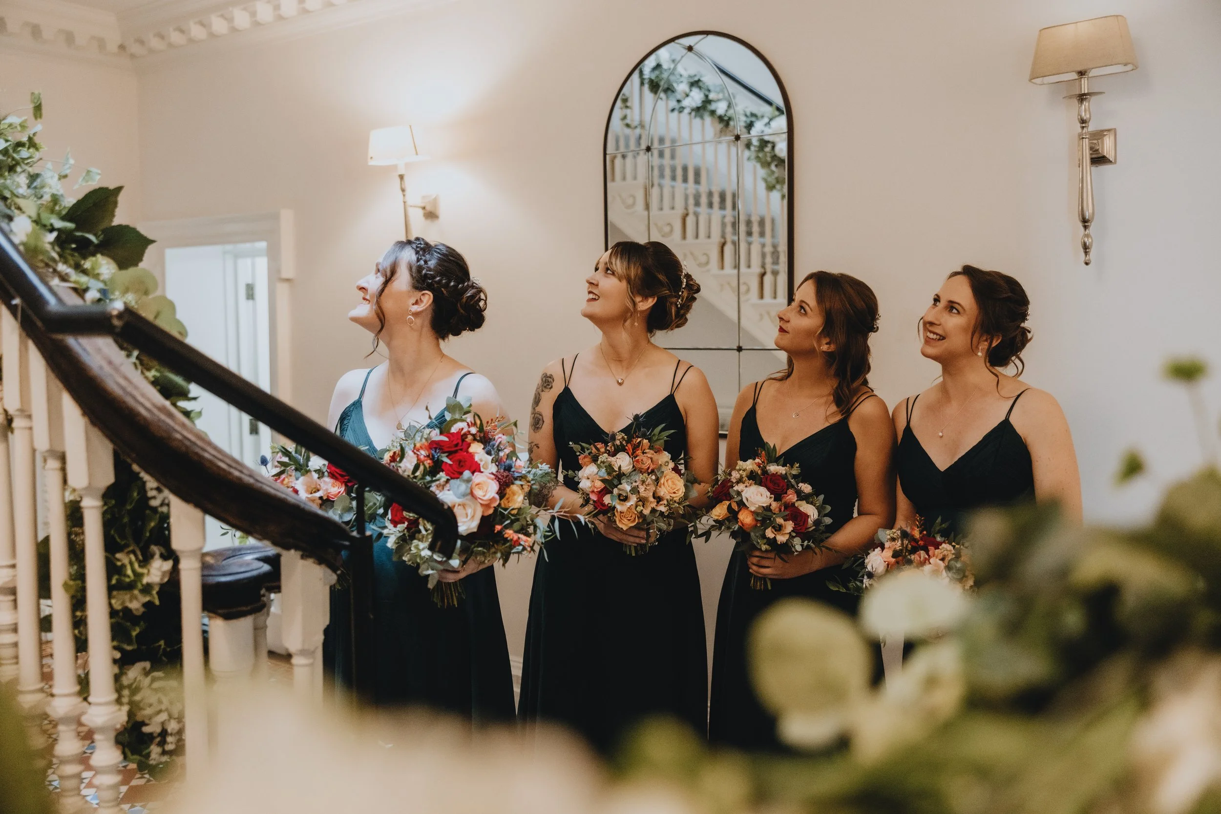 Four women in black dresses holding bouquets of flowers standing on a staircase inside a decorated home. Rowton castle wedding venue 