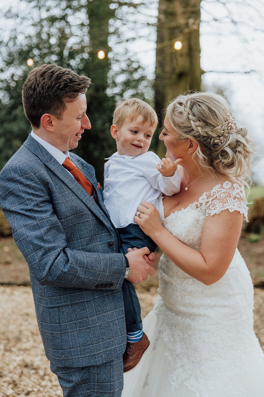 Bride and groom holding a young boy, smiling and looking at each other outdoors, with trees and string lights in the background.