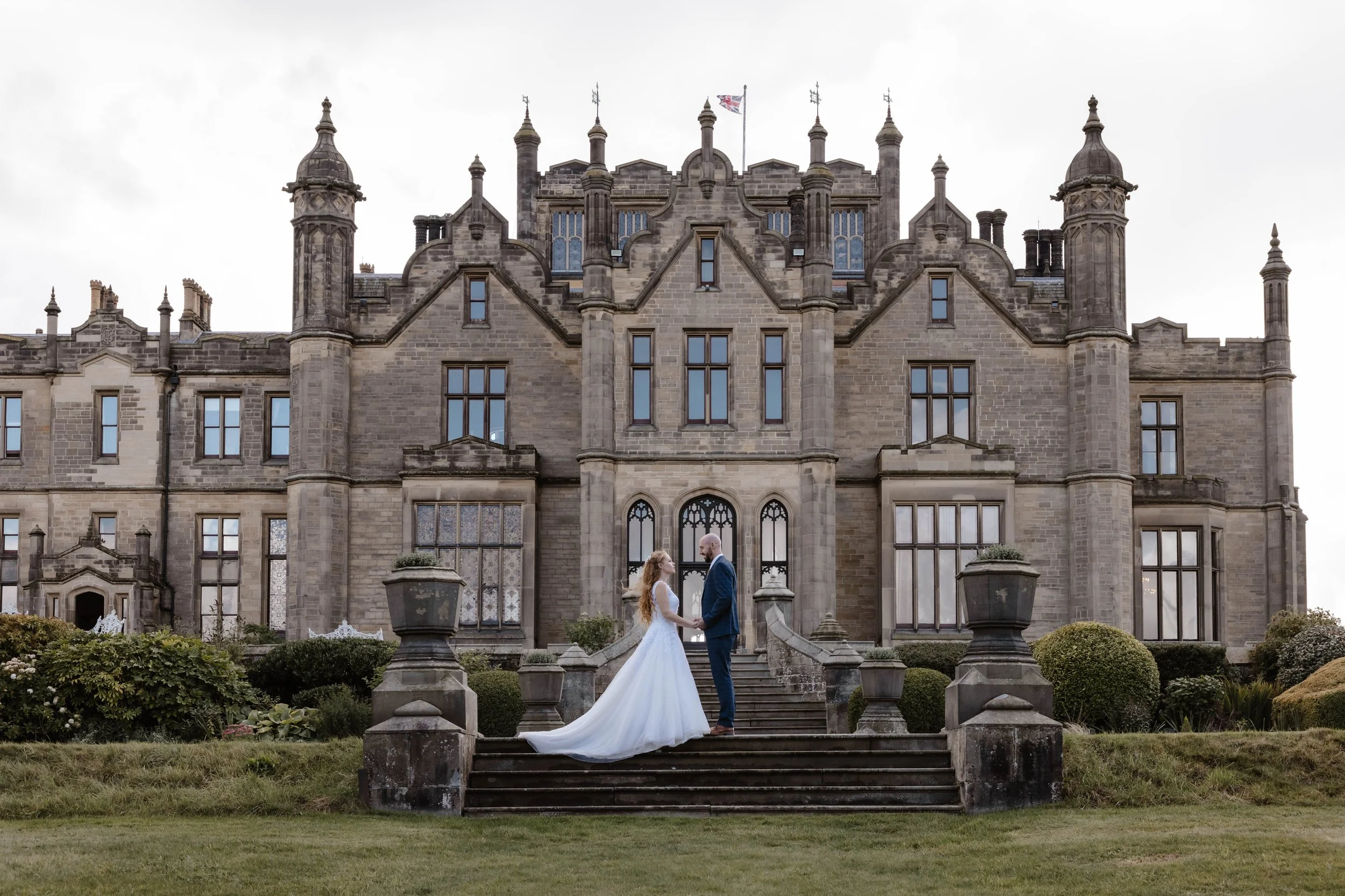 A bride and groom holding hands on the steps in front of a large, historic castle-like building with gothic windows. peckforton castle 