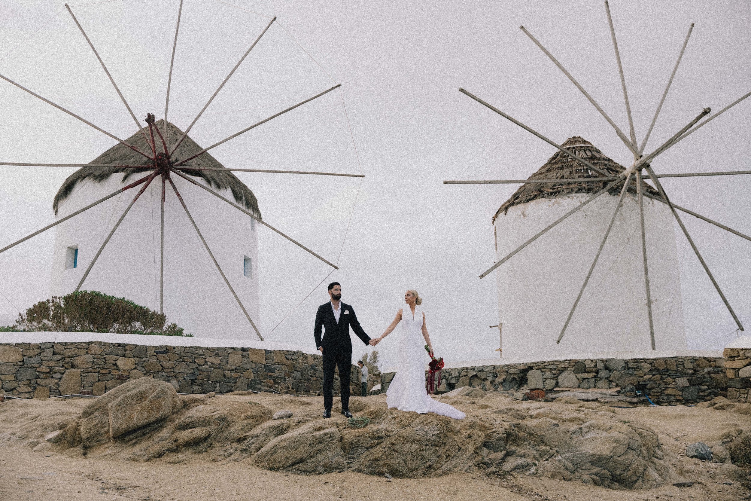 A bride and groom holding hands on a rocky beach with two windmills and umbrellas in the background.