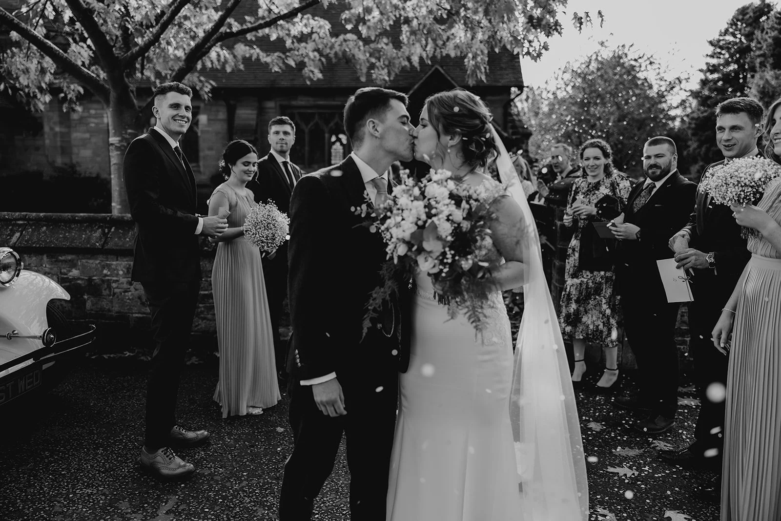 A black and white photo of a wedding ceremony with a bride and groom kissing, surrounded by guests and holding bouquets, outdoors near a church and a vintage car. wedding  merrydale manor 