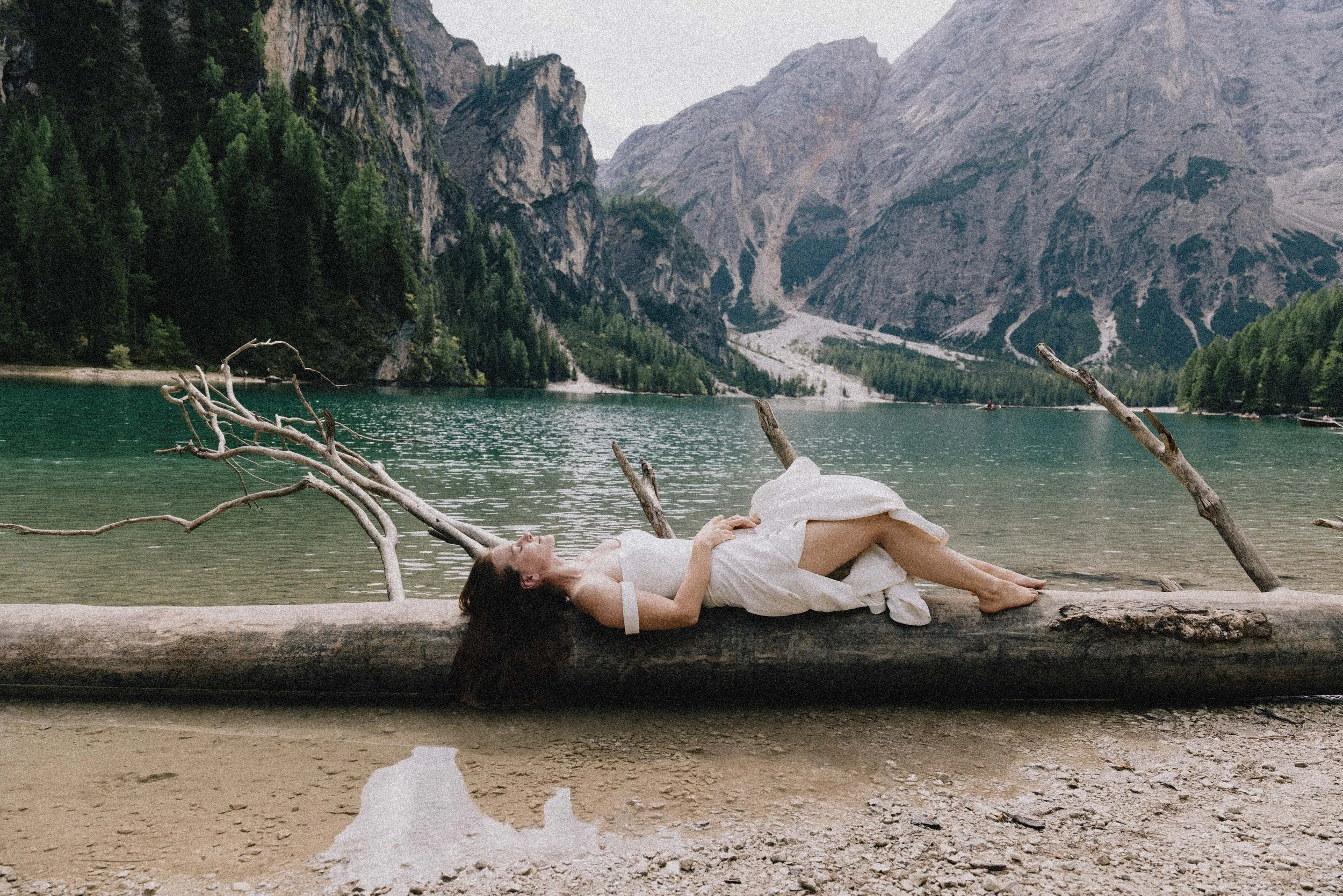 A woman lying on a fallen tree trunk by a lake with mountainous scenery and trees in the background.