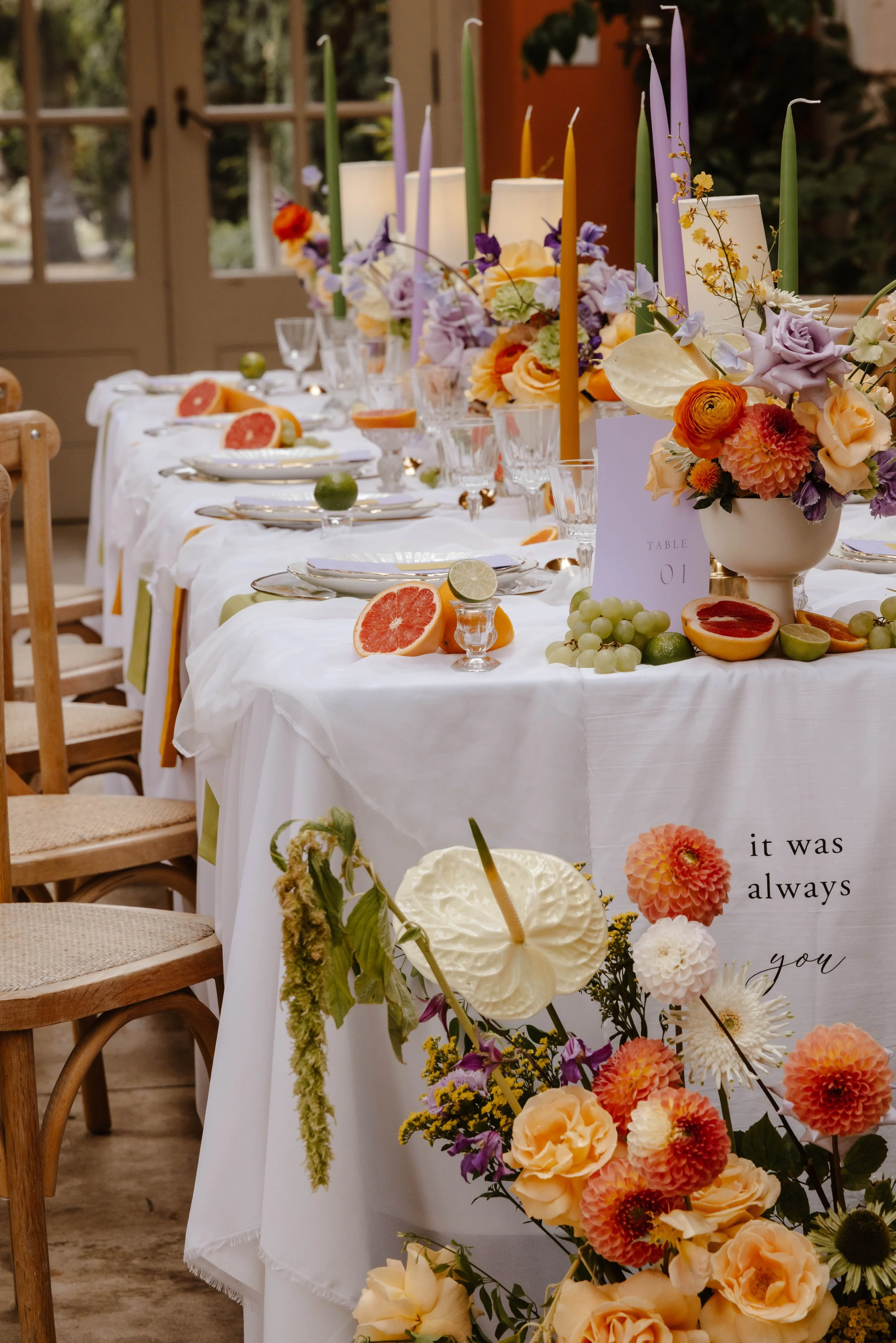 A long banquet table decorated with floral arrangements, candles, and assorted fruit including grapefruit, lime, and grapes, set in a rustic indoor setting. set up at euridge manor. editoral wedding photographer 