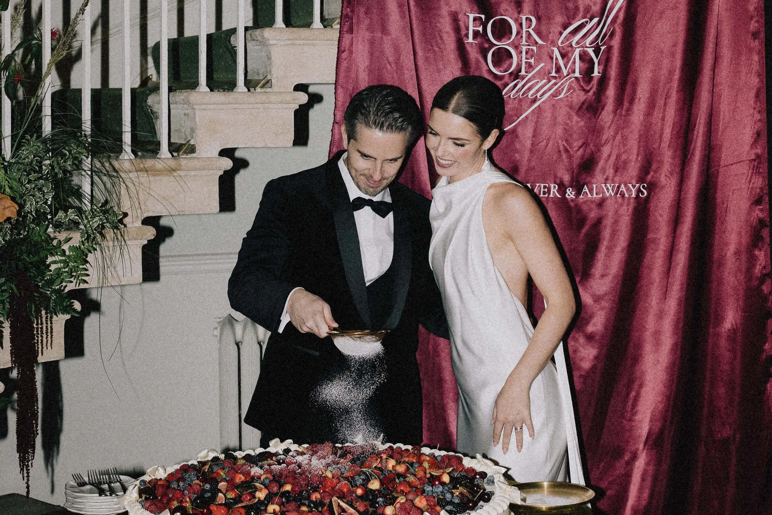 A man in a tuxedo and a woman in a white dress are cutting a large anniversary cake with berries on top. They are standing in front of a red curtain with the text 'For all of my days'. The man is sprinkling powdered sugar onto the cake. middle lodge 