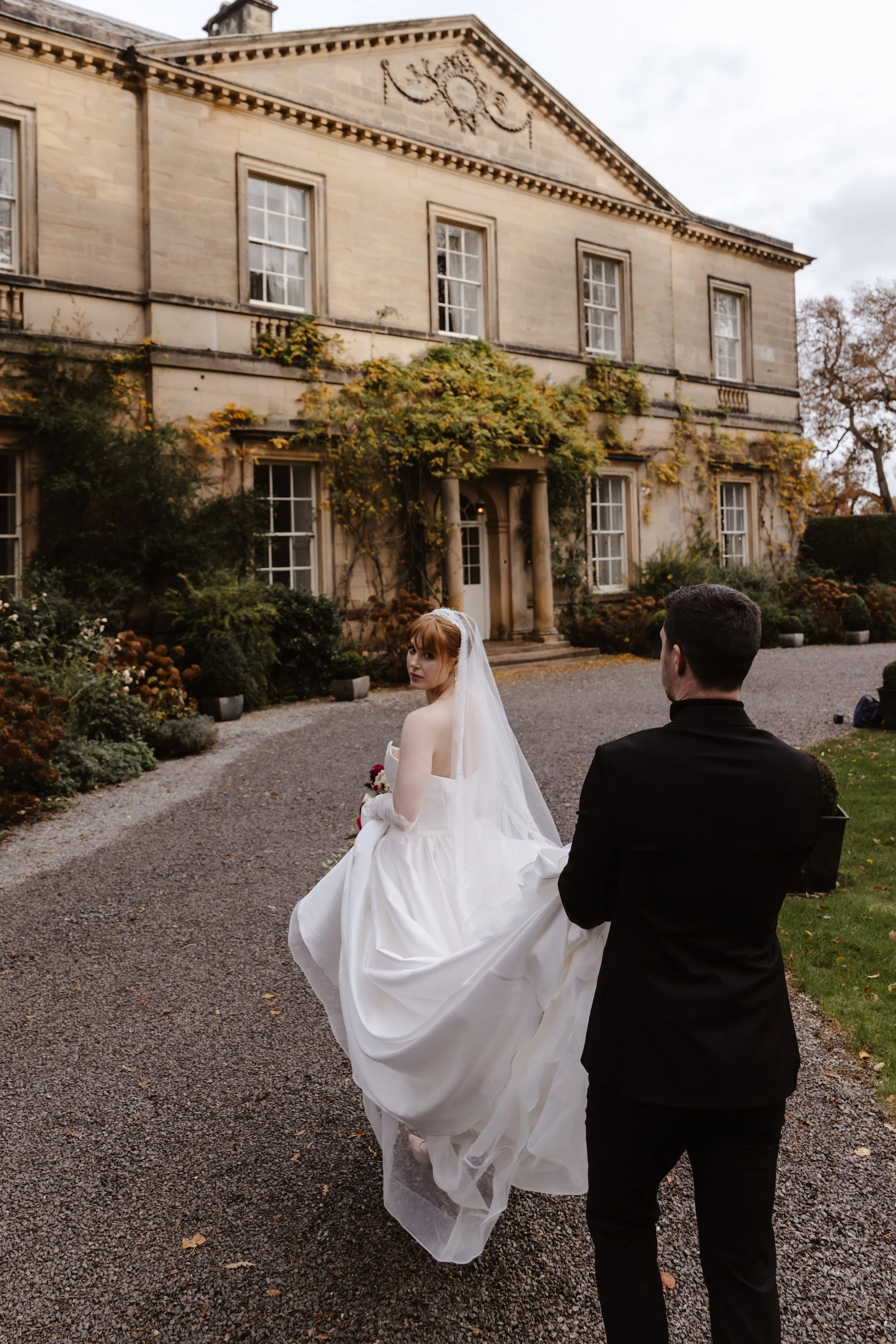 Bride in a white wedding dress and veil walking on a gravel path outside an elegant, historic mansion with vines and foliage, accompanied by a man in a black tuxedo. wedding at middleton lodge, documentary wedding photography