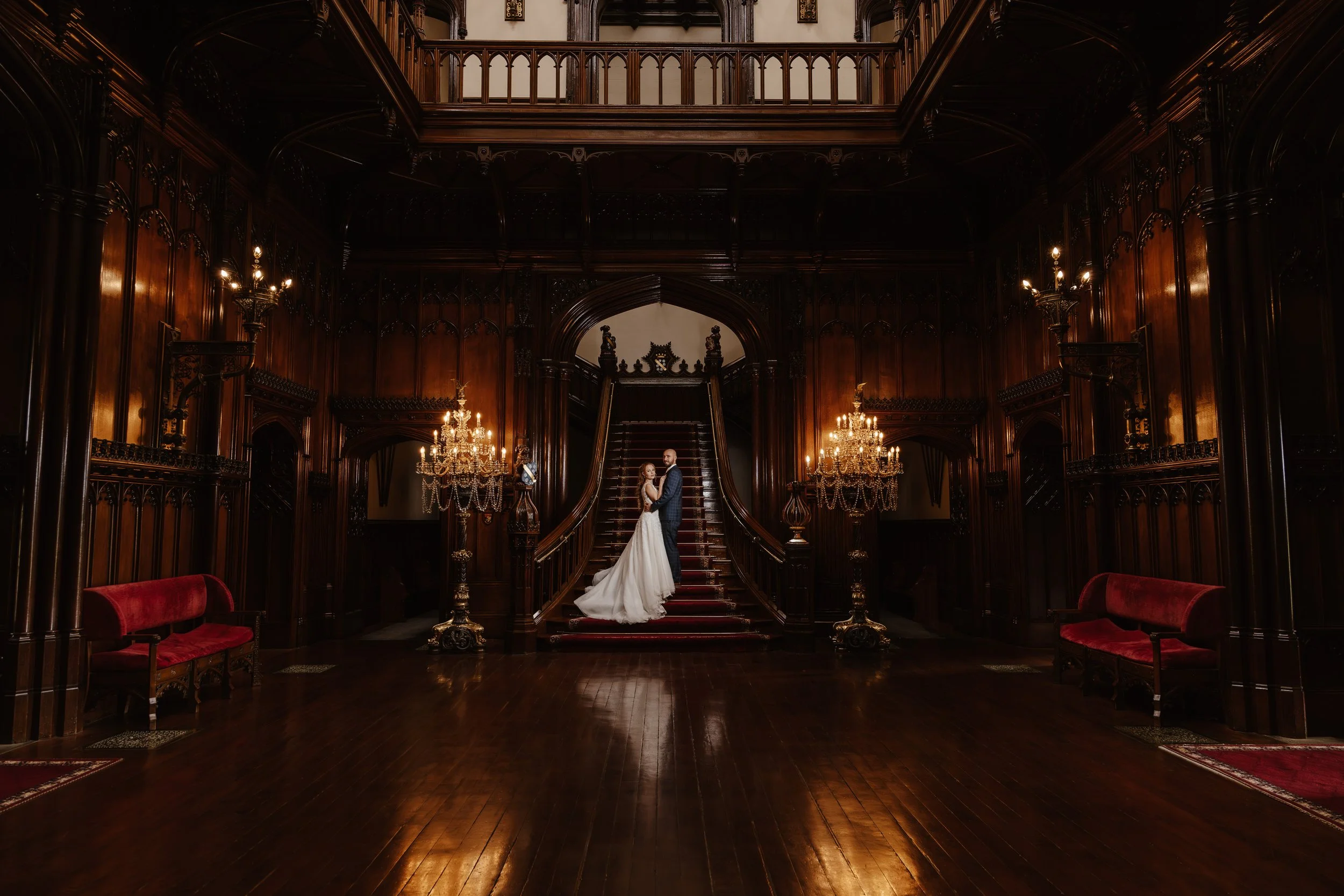 A bride and groom standing on a grand wooden staircase inside an ornate, dark wood-paneled hall with chandeliers and vintage furniture. castle weddings uk 