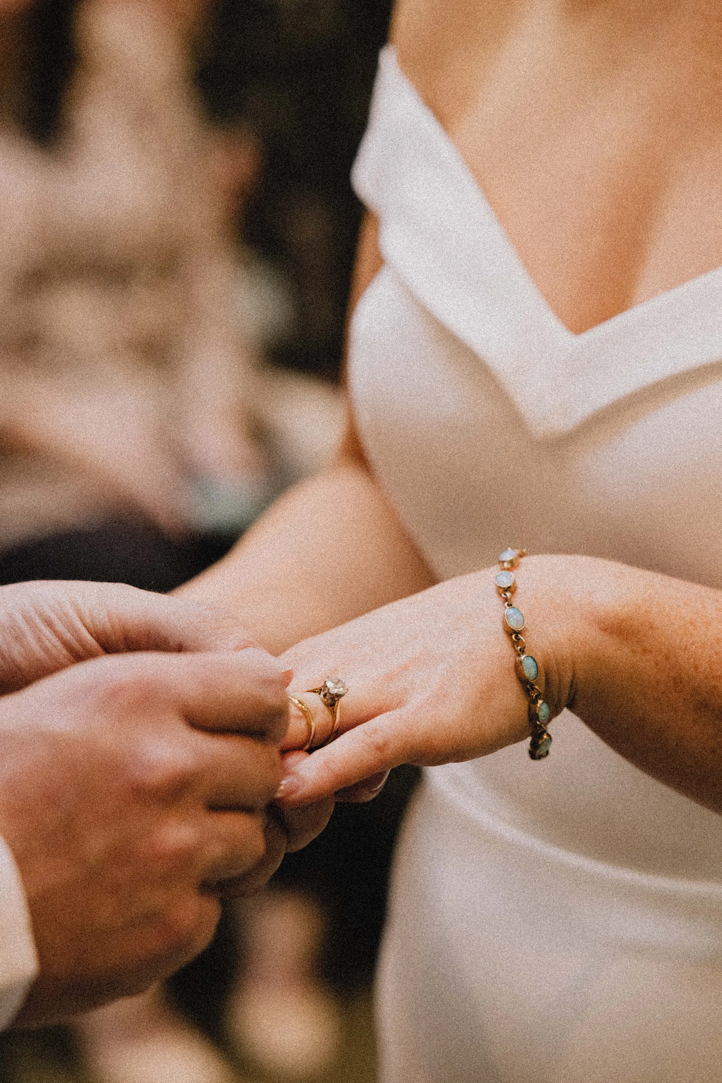 A person wearing a white dress is placing a ring on another person's finger during a wedding ceremony. The person receiving the ring is also wearing a bracelet with oval-shaped stones.