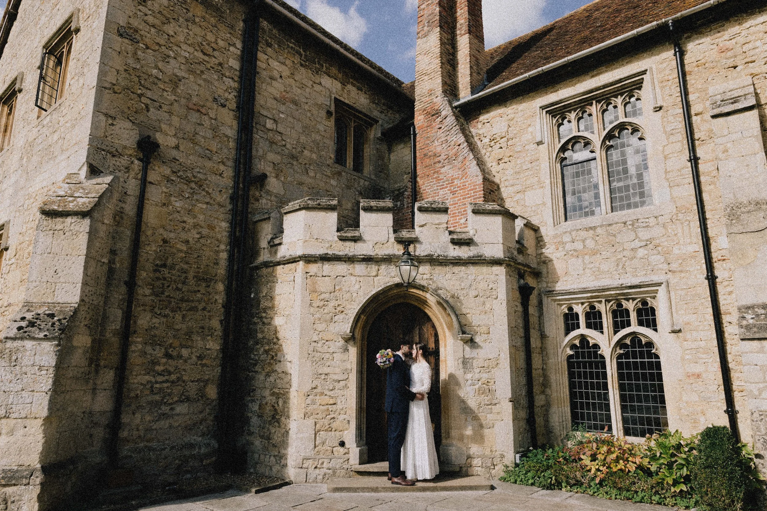 A bride and groom standing close at the entrance of a historic stone building, likely a church, on a sunny day.