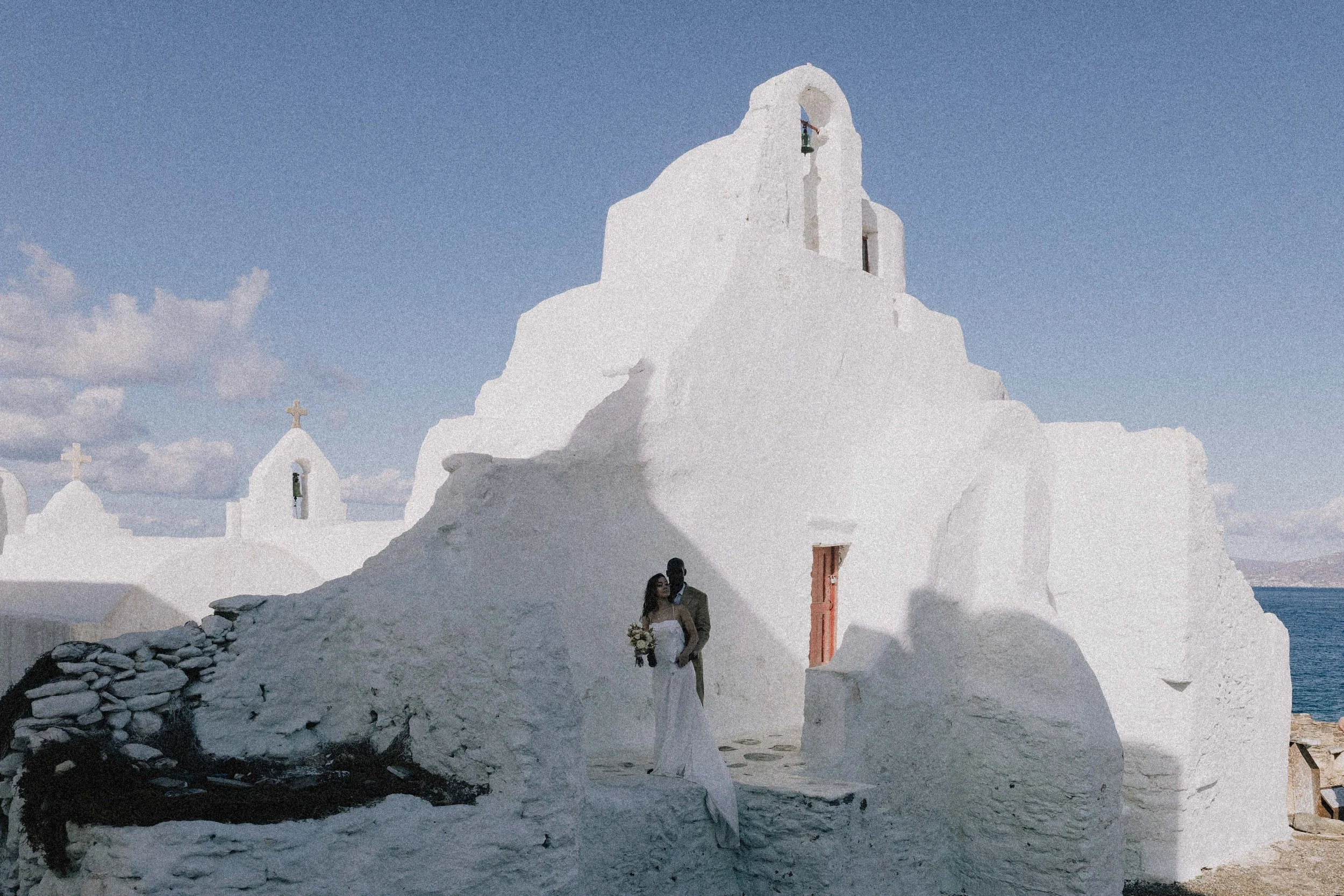A wedding couple standing in front of a whitewashed church with a bell tower, near the sea on a sunny day.
