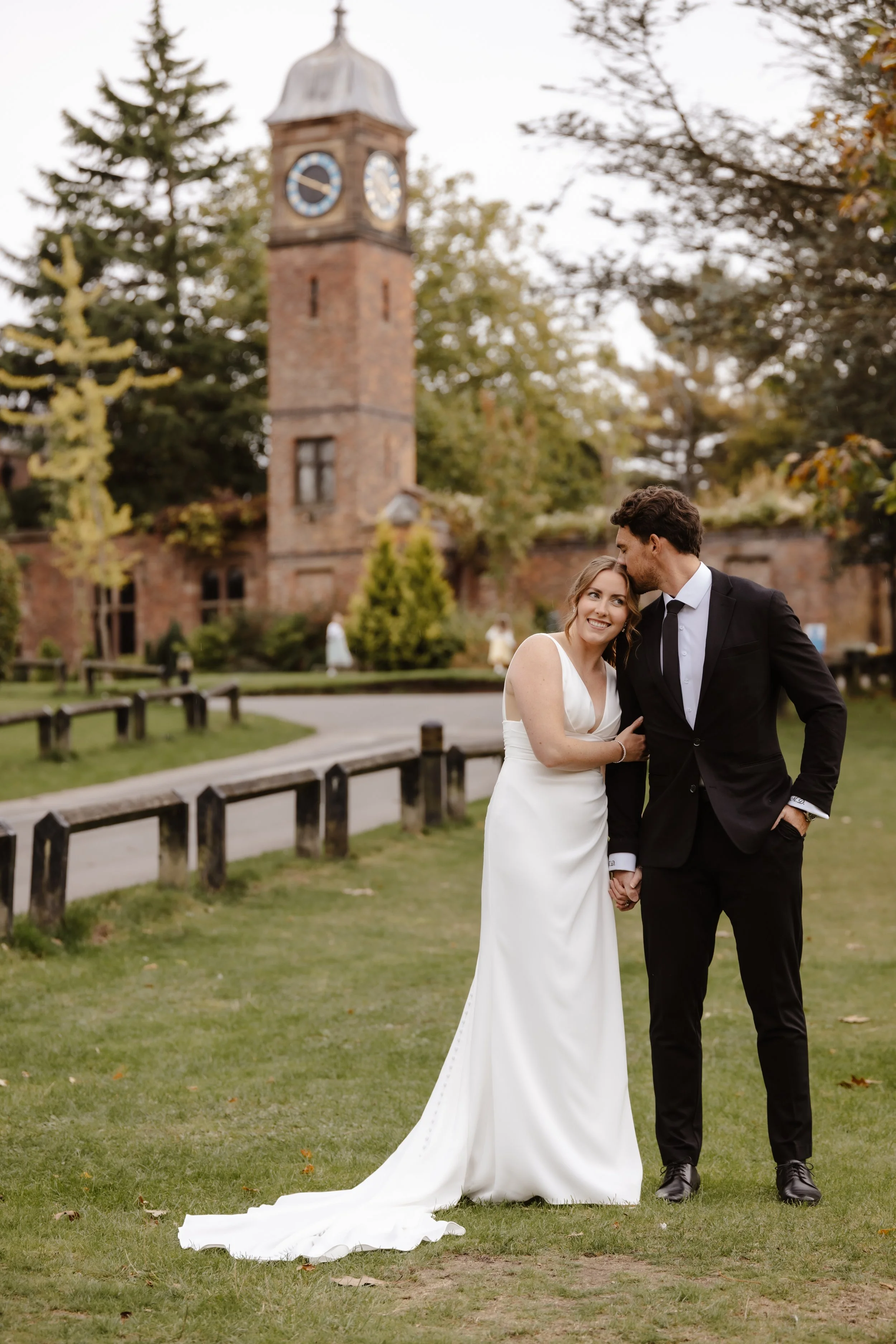 A newlywed couple dressed in wedding attire stands on a grassy area, holding hands, with the bride smiling while the groom kisses her on the forehead. wedding at walton hall gardens in cheshire, documentary style wedding photography