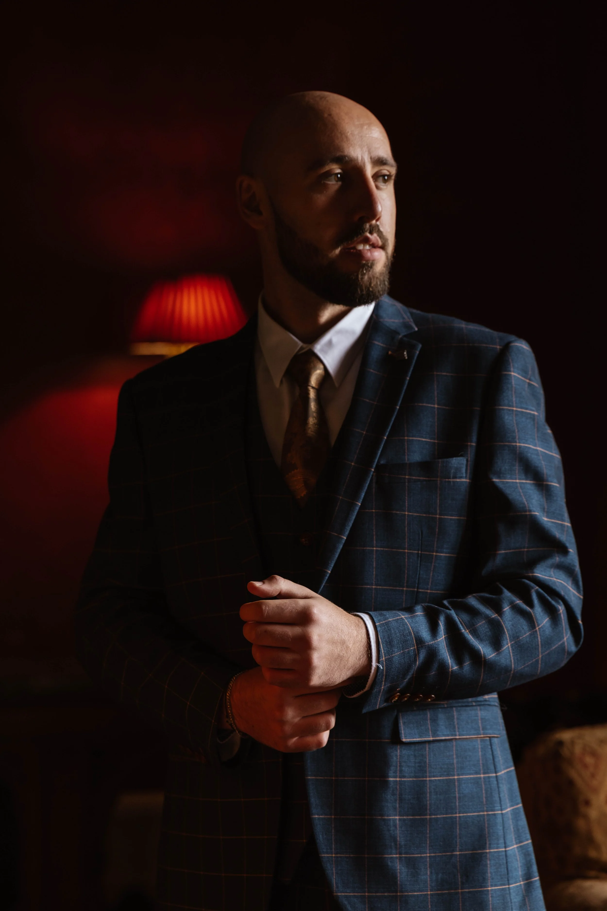 A man with a beard and a shaven head wearing a dark checkered suit, white shirt, and patterned tie, standing in a dimly lit room with a red lamp in the background, looking to the side. styal lodge wedding, groom preparations 