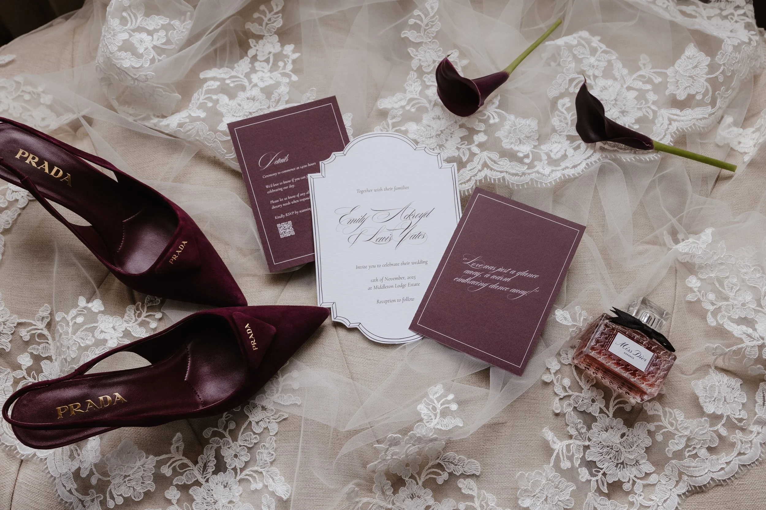 Wedding invitation suite with burgundy accents, Prada shoes, dark purple calla lilies, lace fabric, and Miss Dior perfume on a lace tablecloth. editorial shot of details at middleton lodge 