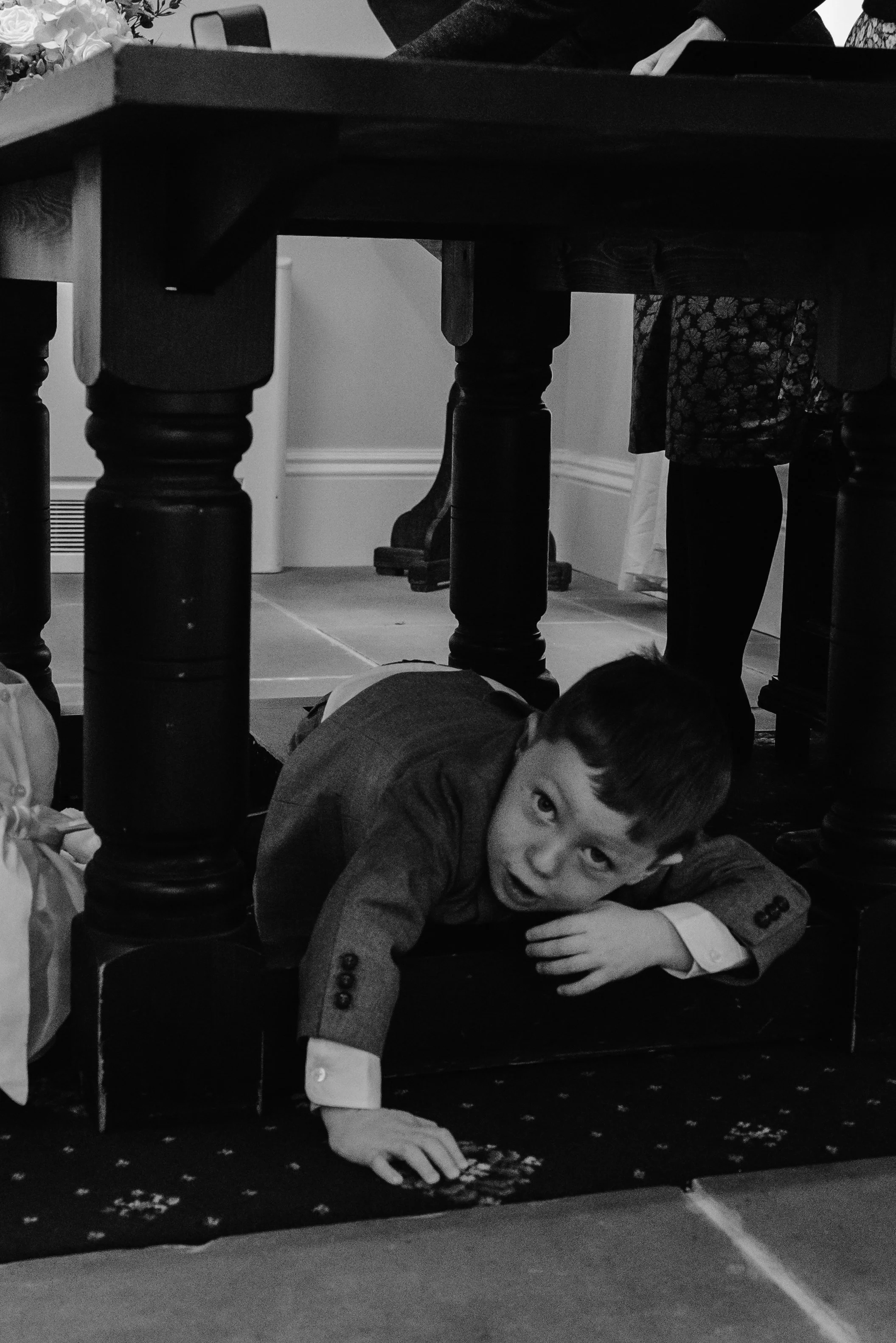 A young boy in a suit is crawling under a dining table with wooden legs. The boy is looking at the camera with an expression of surprise or curiosity. The image is in black and white. York registry office wedding 