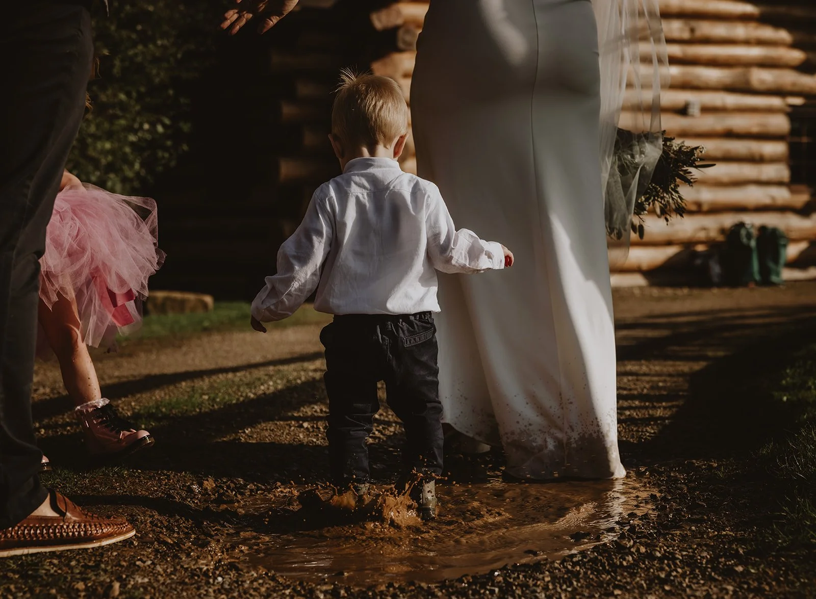 A young boy in a white shirt and black pants walking through a muddy puddle at an outdoor event, with adults and children around. at uk wedding venue hidden river cabins 