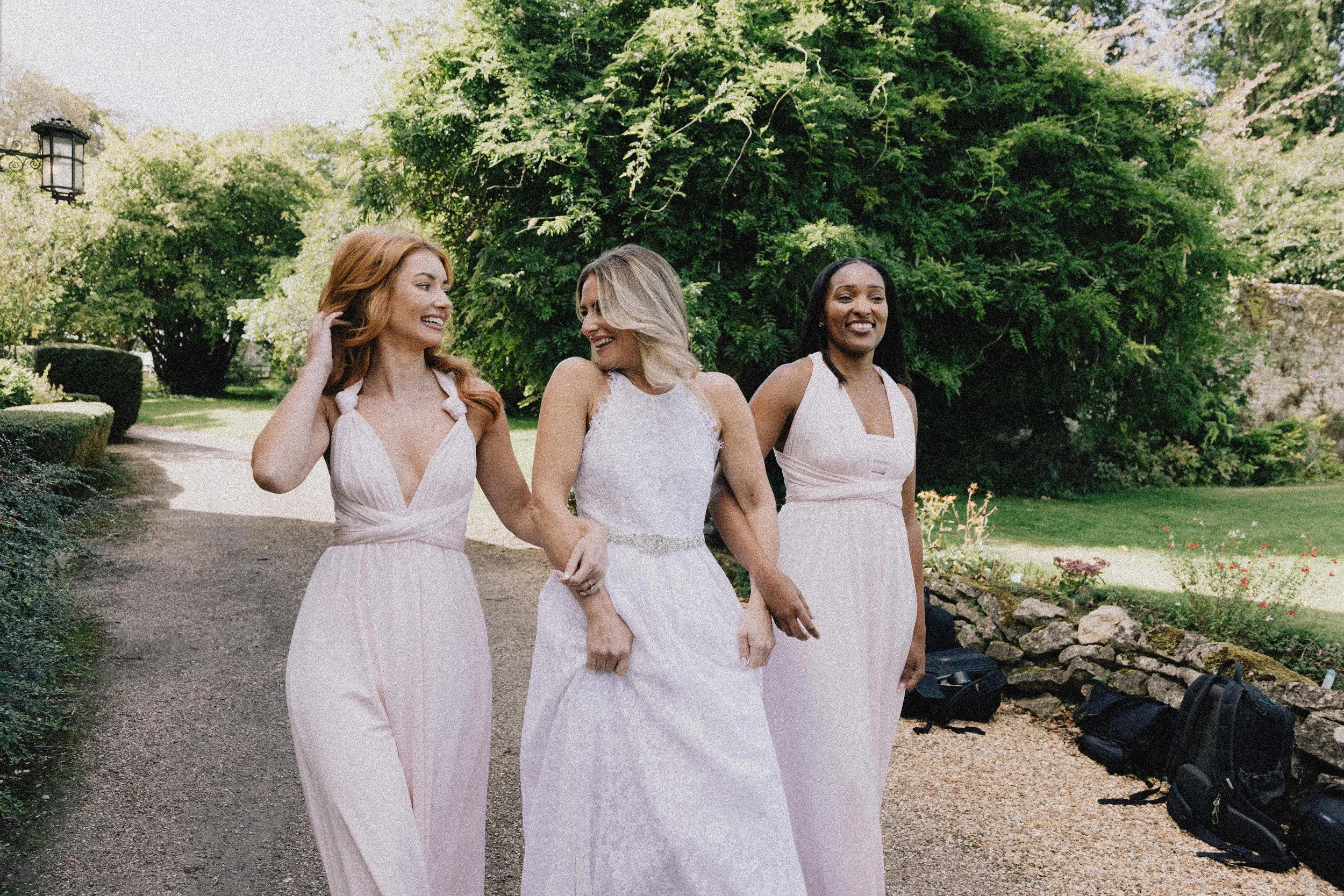 Three women in light pink dresses walking outdoors on a garden path, smiling and laughing, with lush green trees in the background.