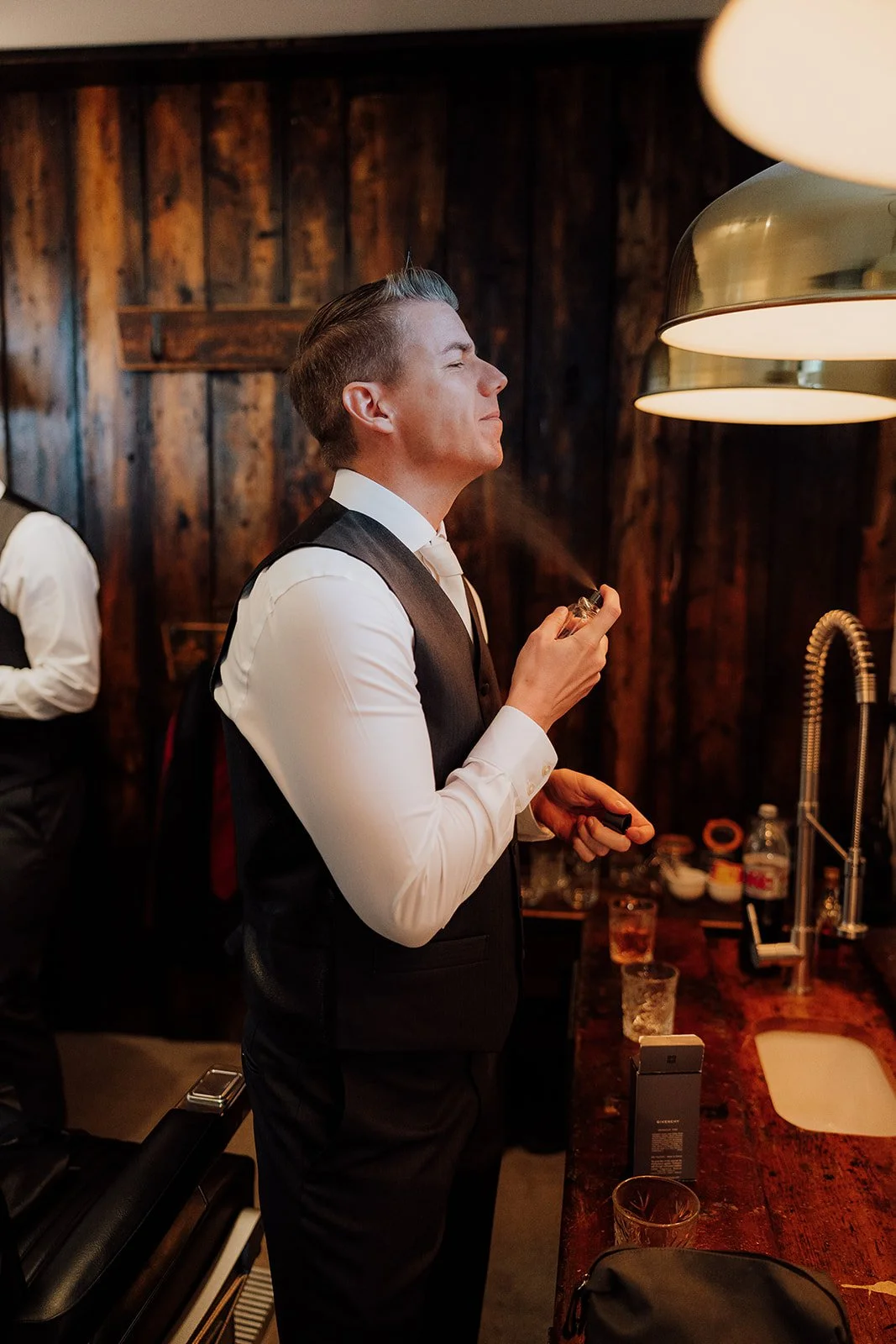 A man in a black vest and white shirt stands at a bar, holding a perfume bottle and opening it. The background features a wooden wall with warm lighting, and a man in similar attire is visible on the left side. peckforton castle venue