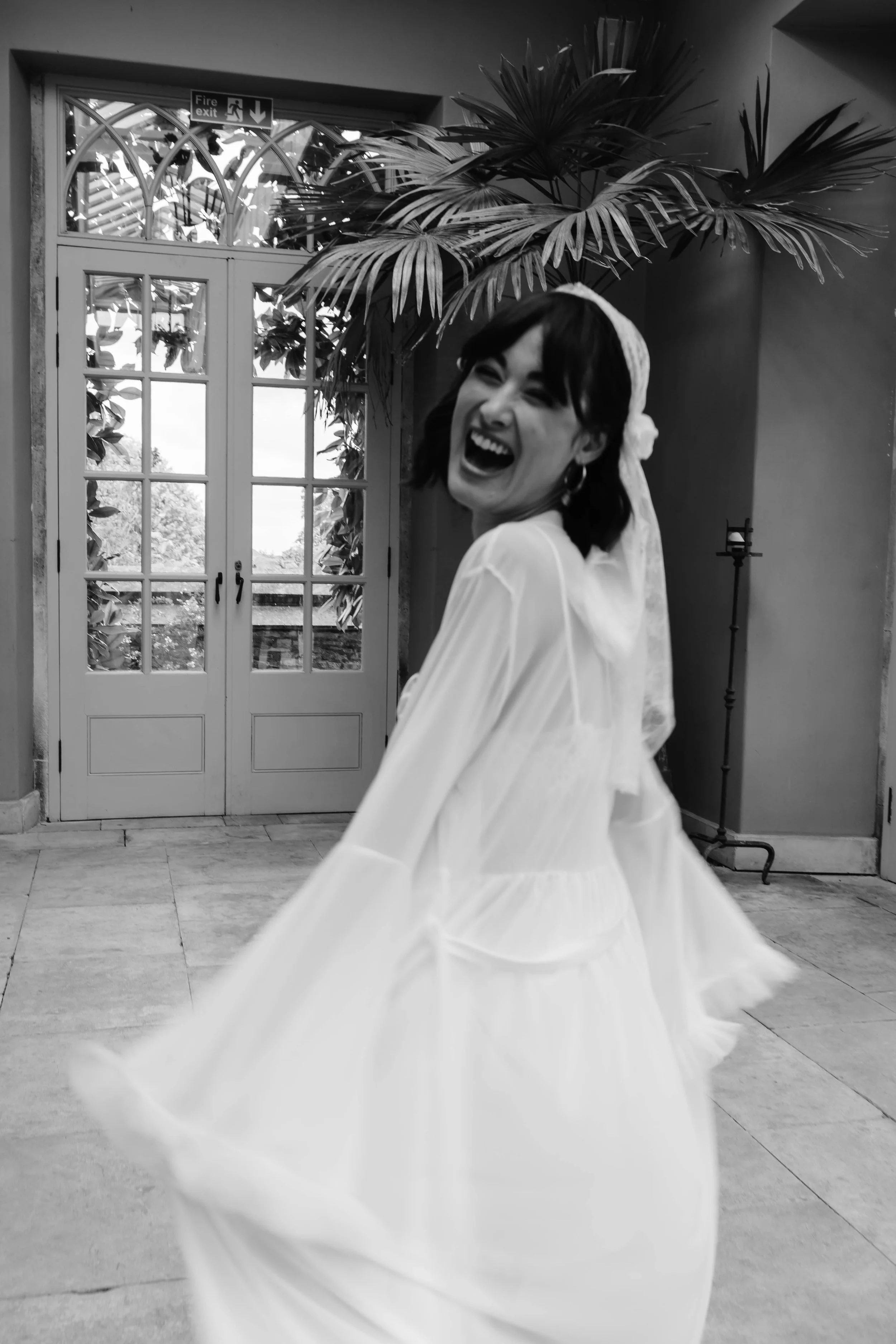 Woman in a wedding dress laughing indoors with large potted plants and glass doors in the background. wedding arley hall in cheshire