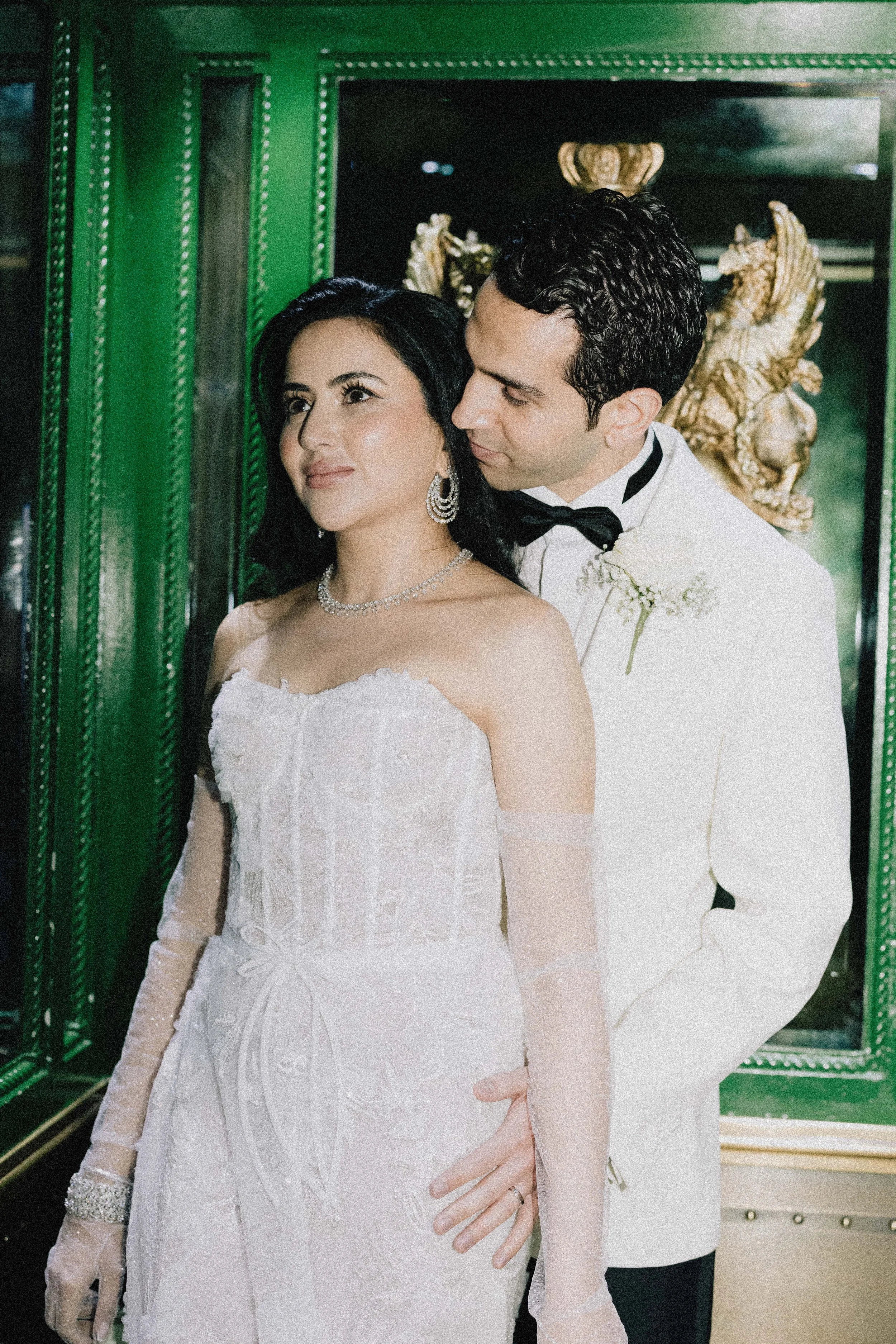 A bride and groom in wedding attire pose together indoors with ornate green and gold decor.