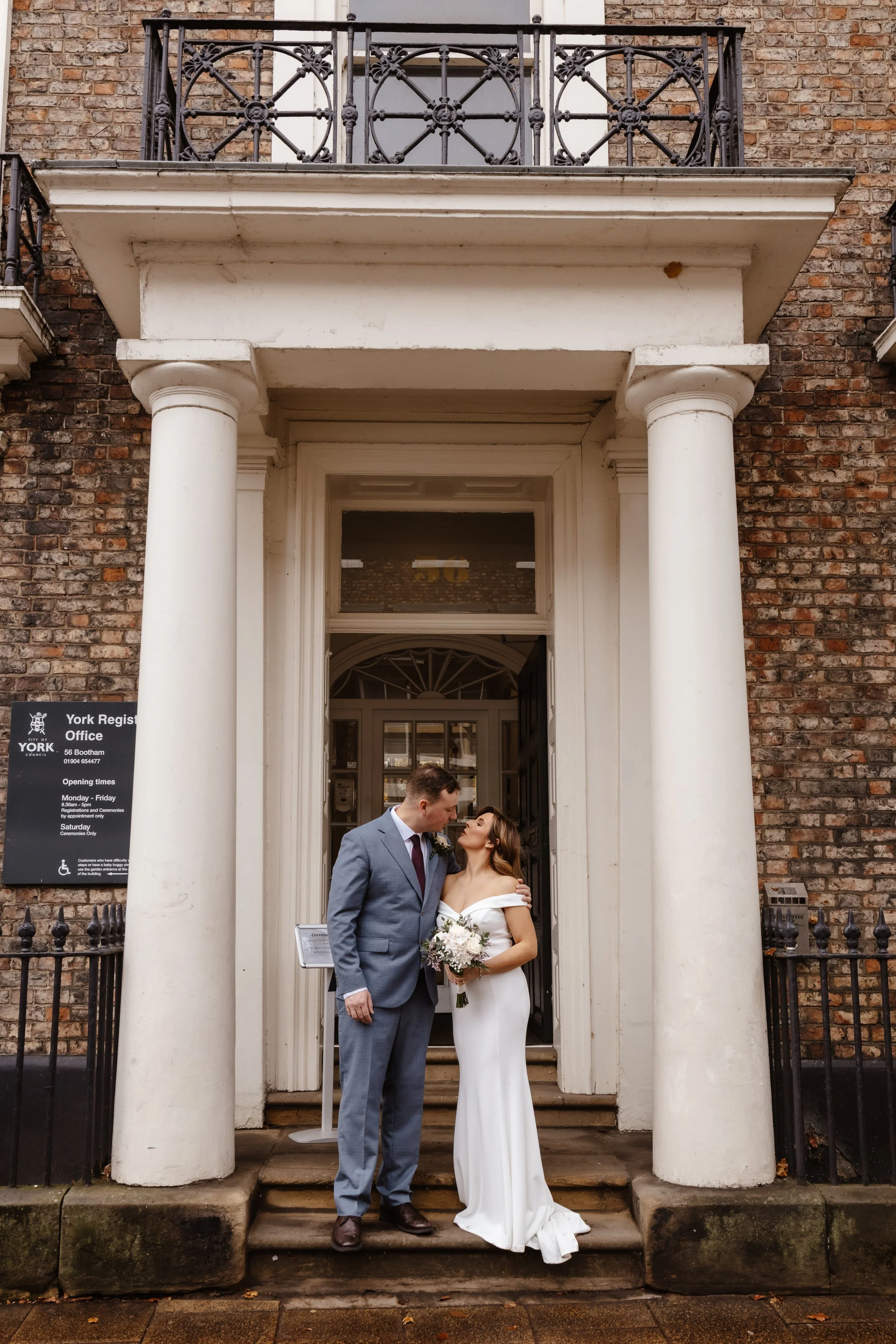 A bride and groom standing on steps outside a building, sharing a moment. The groom is in a gray suit, and the bride is in a white wedding dress holding a bouquet. The building has brick walls and white columns. York registry office wedding 