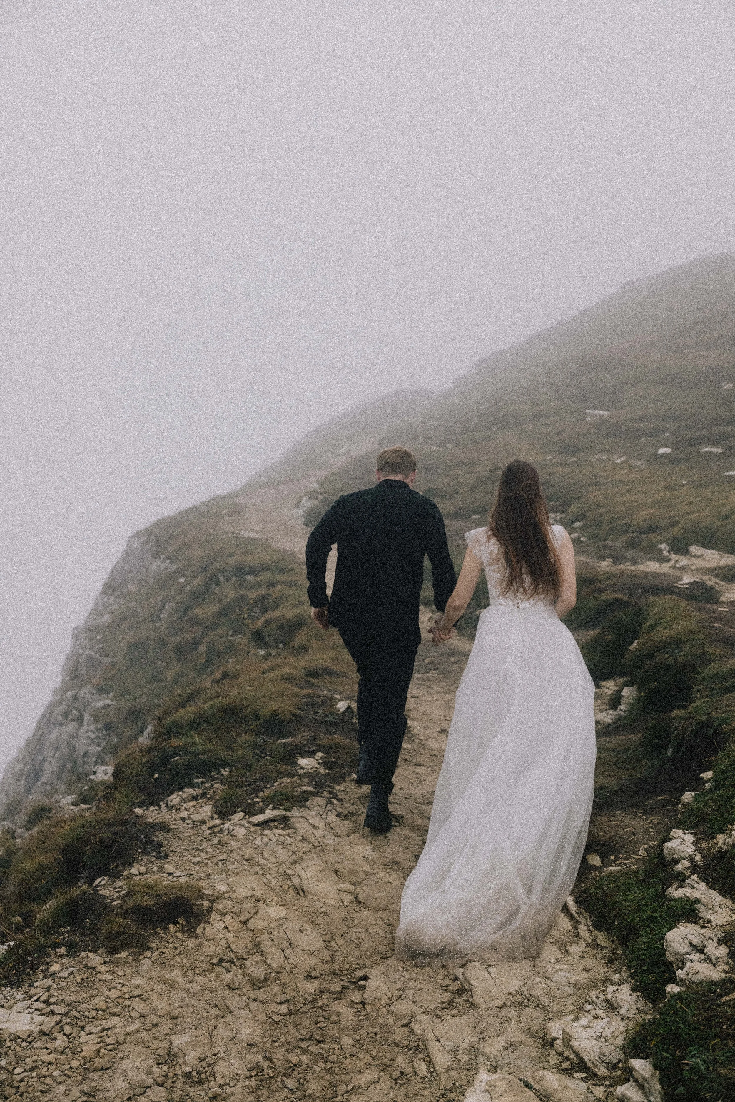 A couple, a man in a black suit and a woman in a white dress, holding hands and walking along a narrow mountain trail surrounded by fog and mist.