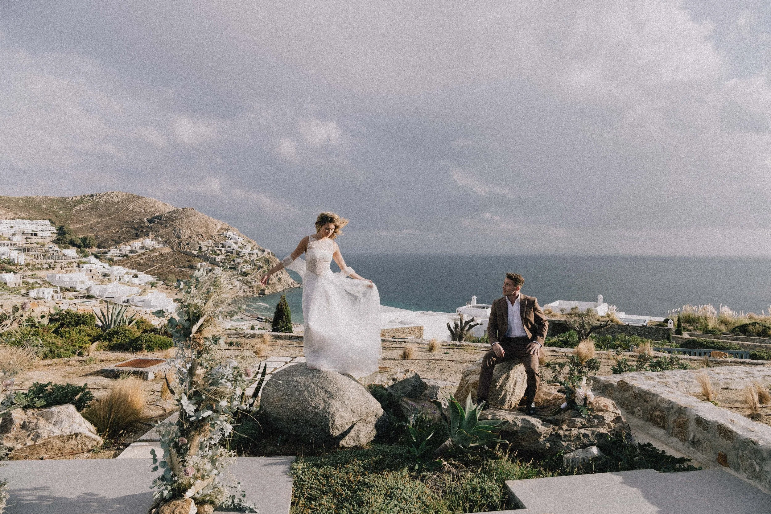 A bride in a white wedding dress standing on a large rock with her hand holding the edge of her dress, while a groom in a brown suit sits on another rock looking at her, with a scenic coastal landscape and cloudy sky in the background. greece wedding