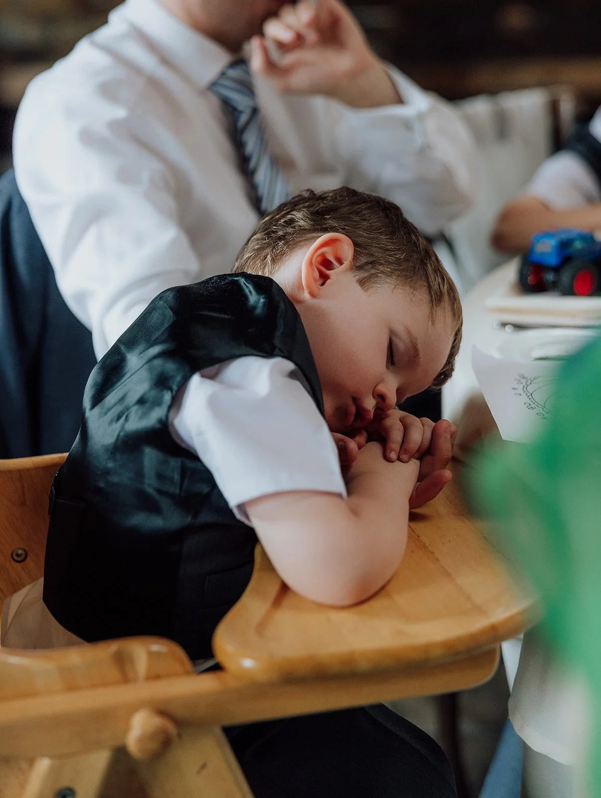 Young boy sleeping on a wooden high chair at a table, wearing a white shirt and black vest, with an adult man in business attire sitting nearby. Merrydale Manor Wedding Venue
