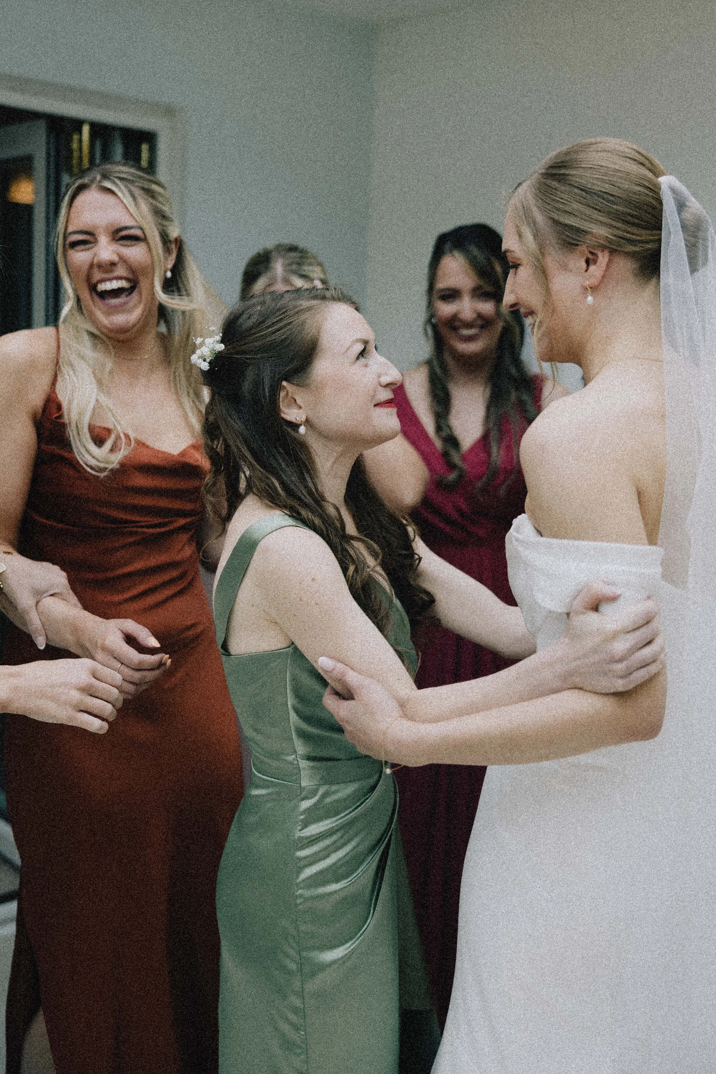 A group of women, including a bride in a white wedding dress, sharing an emotional moment with a woman in a green dress, surrounded by friends in elegant dresses, at a wedding celebration.