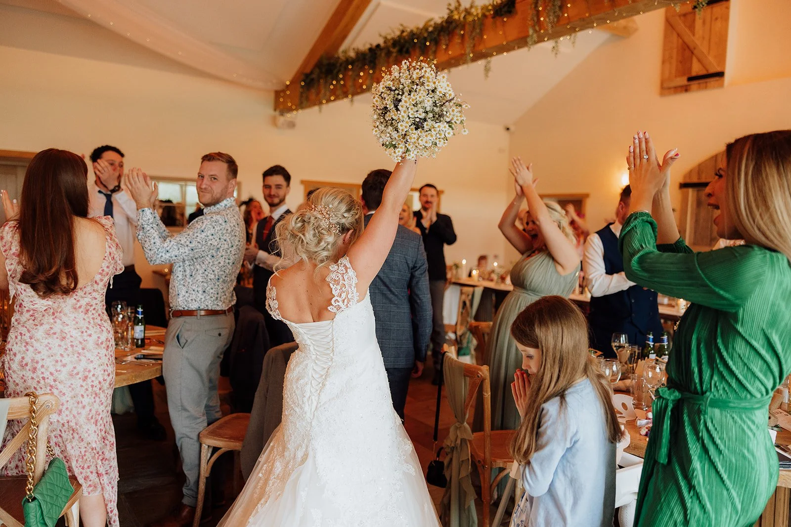 A bride in a white wedding dress holding a bouquet of flowers above her head, celebrating at her wedding reception with guests clapping and smiling in a warmly decorated indoor venue. Sandhole Oak Barn wedding. venue, documentary wedding photography