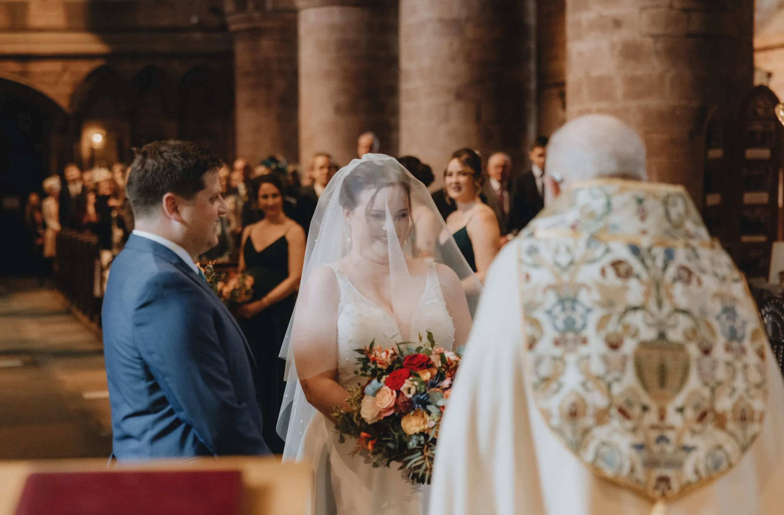 A bride and groom standing at the altar during a wedding ceremony in a church, with a priest officiating. The bride is wearing a white wedding gown with a veil and holding a colorful bouquet. The groom is in a blue suit. Guests are seated in the back