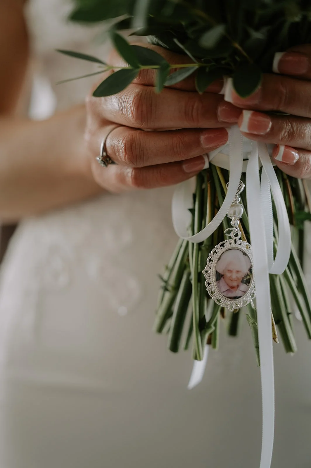 Close-up of hands holding a small bouquet of green leaves, decorated with white ribbons and a framed photo of an elderly woman, with a wedding ring on the ring finger.