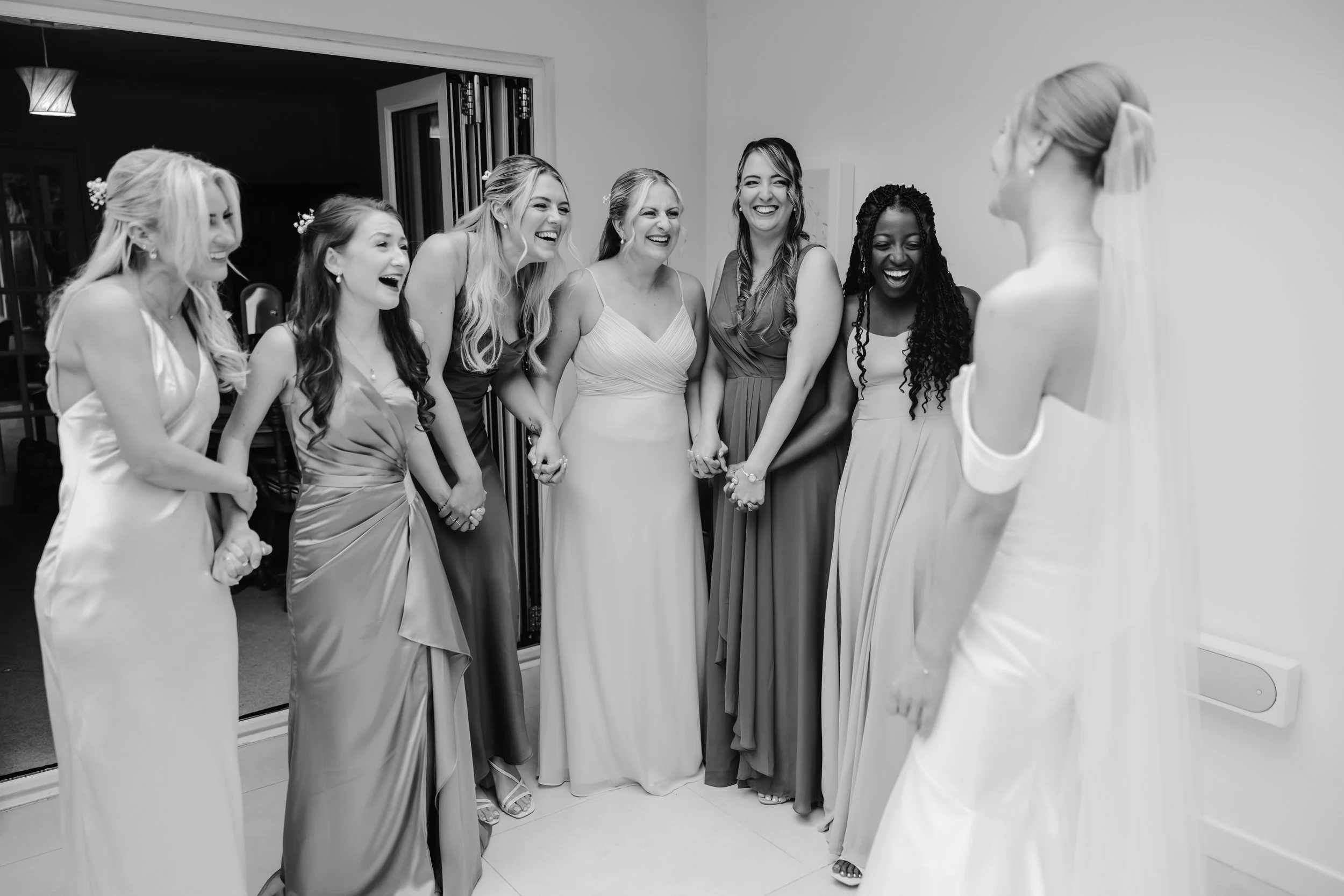 Group of women dressed in formal gowns laughing and holding hands in a room, with a bride on the right side facing them. documentary wedding photographer in a wedding in cheshire