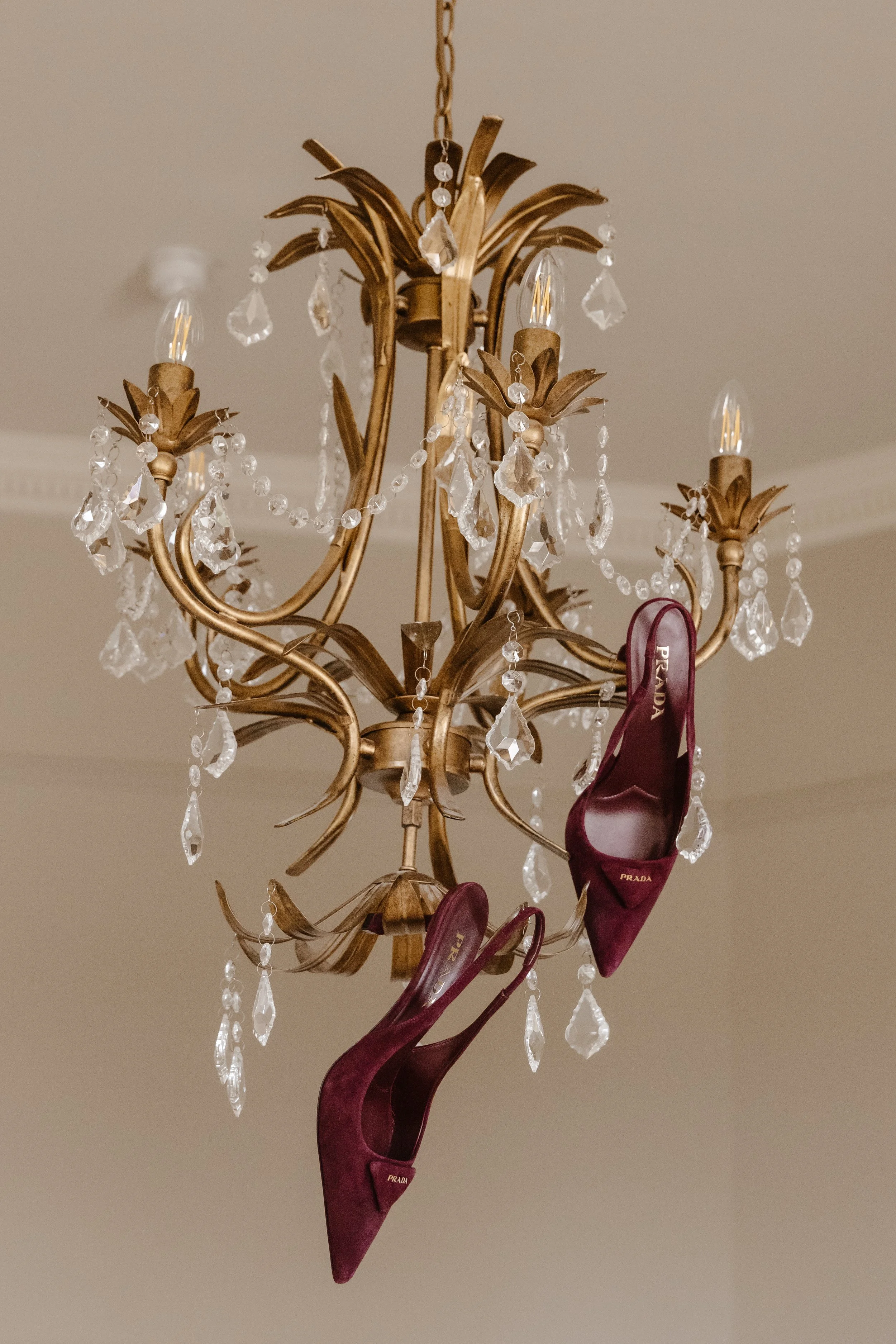A chandelier with crystal adornments hanging from a ceiling, with a pair of burgundy Prada high heel shoes draped over one of the chandelier's arms. Editorial shot as seen in vogue 