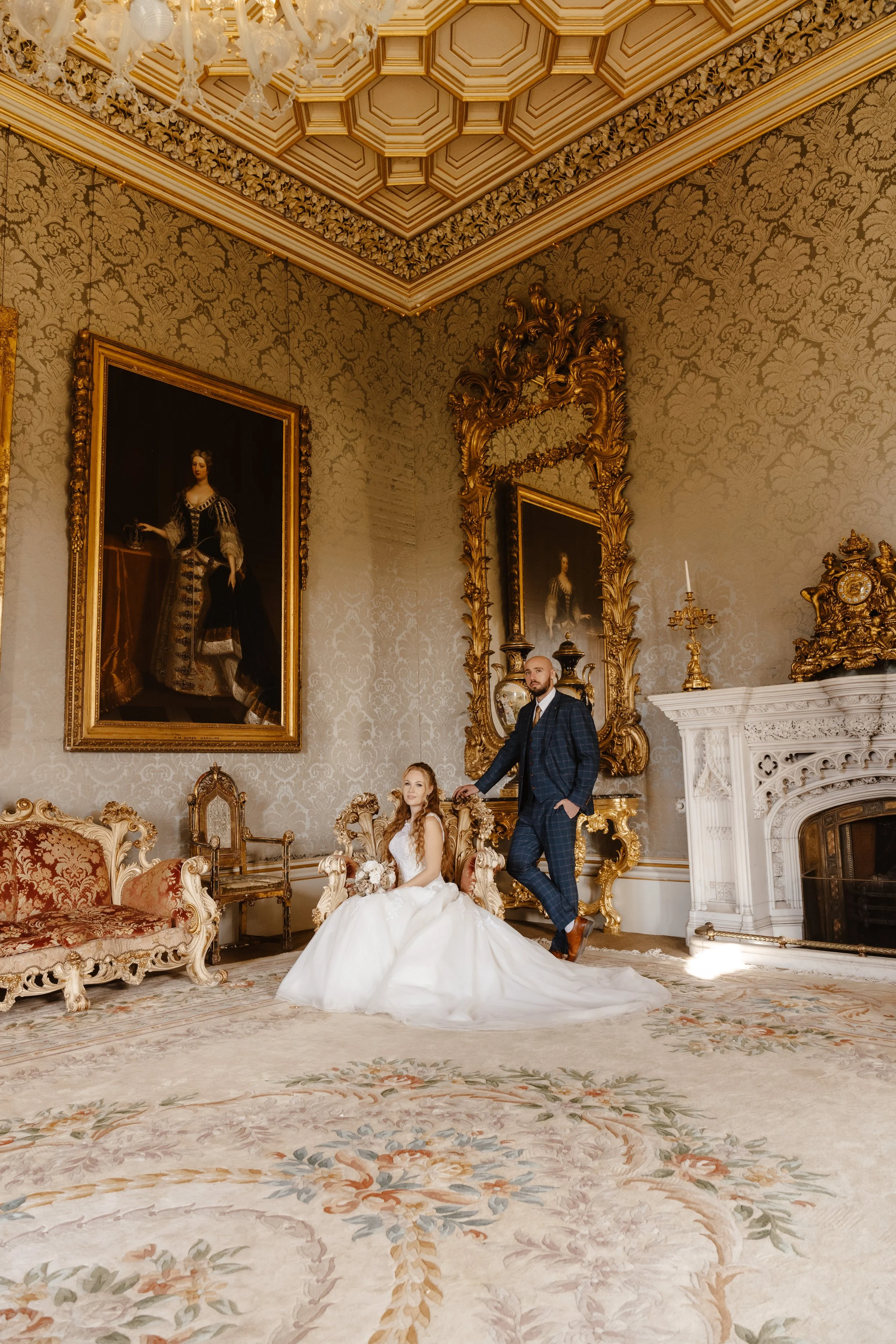 A bride in a white wedding gown and a groom in a blue checkered suit pose in an ornate, gold-accented room with classical artworks, vintage furniture, and a luxurious fireplace. wedding at rowton castle 