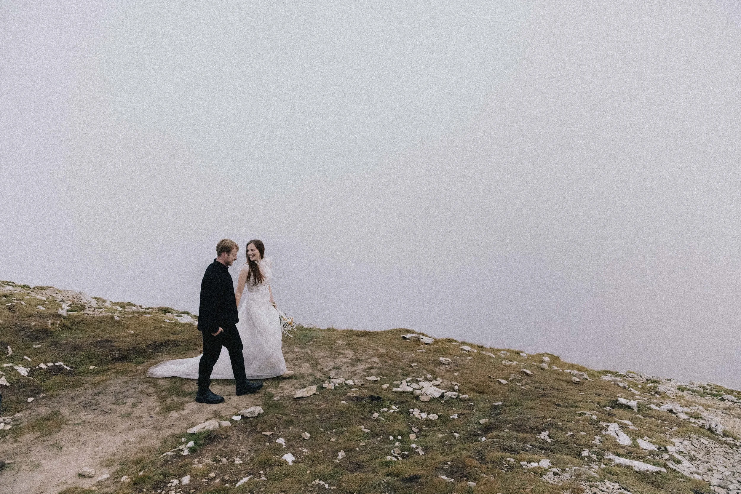 A bride and groom walking together on a rocky, grassy hilltop with a foggy, overcast sky in the background.