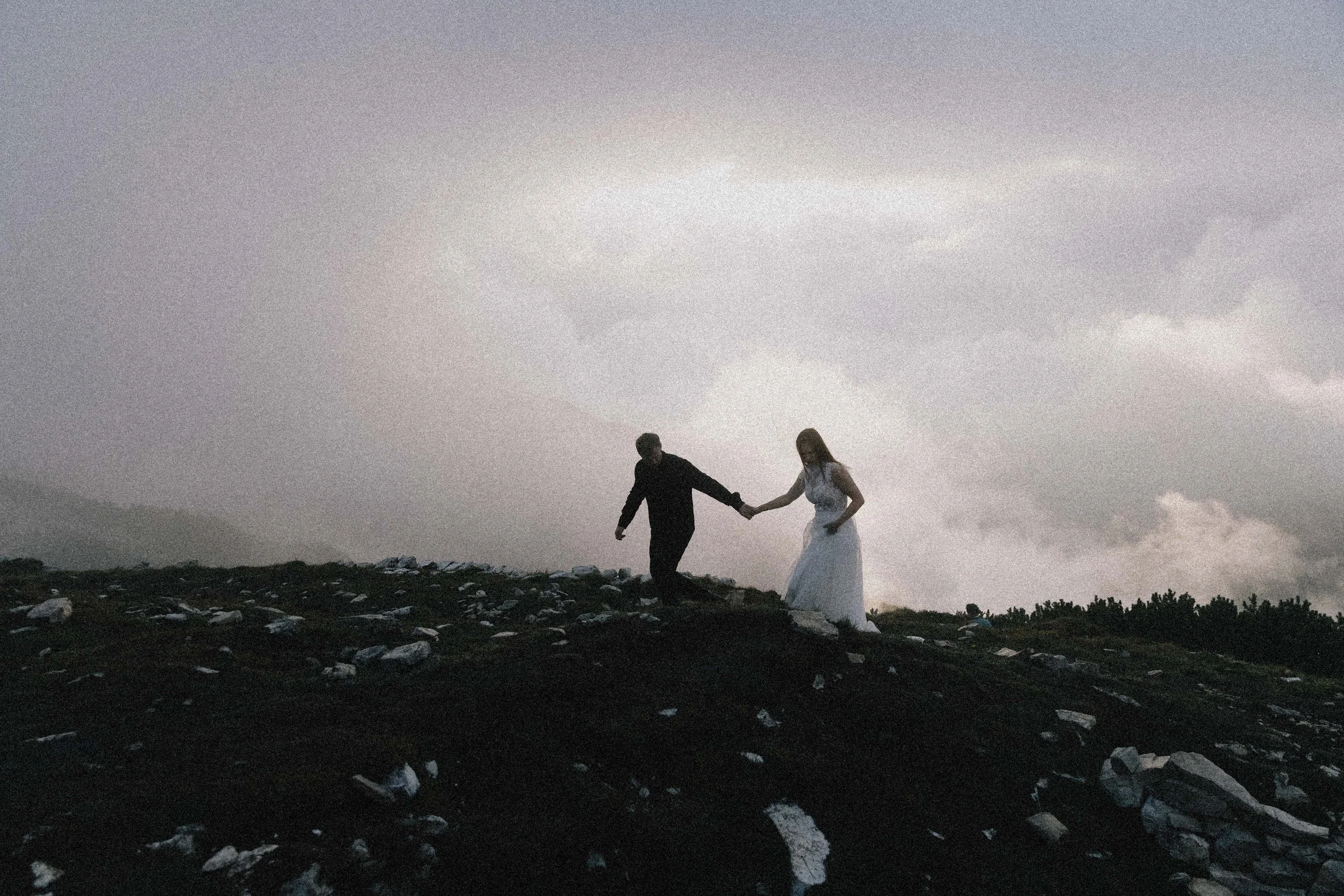A couple dressed in wedding attire holding hands and walking on a rocky hilltop under a cloudy sky.